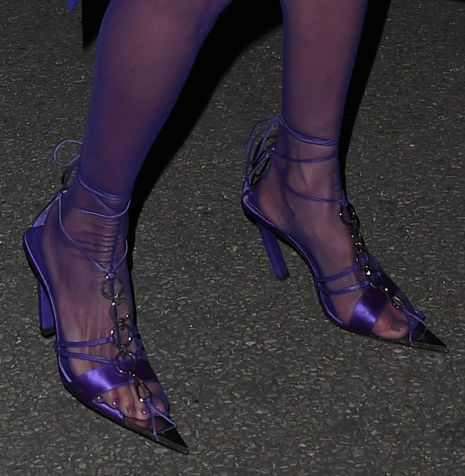 Alessandra Ambrosio finishes her monochrome purple look with satin lace-up heels, featuring sheer panels, pointed toes, and bold metal ring accents that echo the outfit’s sultry edge
