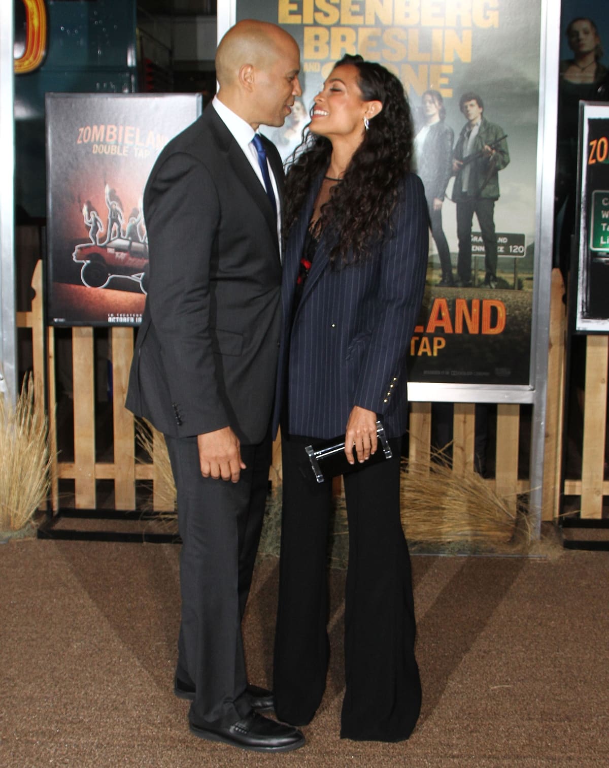 Rosario Dawson pairs wide-leg trousers with platform heels and a sharp pinstripe blazer, standing nearly eye-to-eye with Cory Booker at the Zombieland: Double Tap premiere in Los Angeles