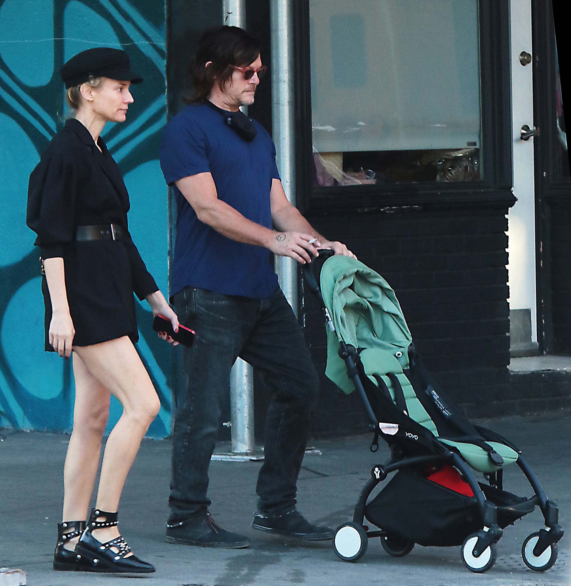Pushing a stroller through Soho, Norman Reedus keeps things laid-back in a blue tee, dark jeans, and black boots, while Diane Kruger contrasts his casual look with a polished black mini dress, belted waist, and studded flat shoes that sharpen her off-duty style