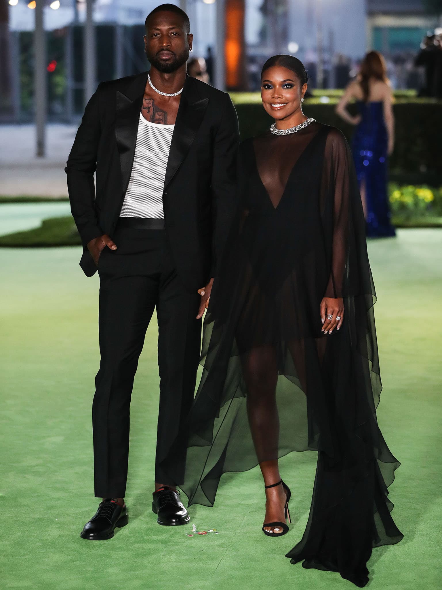 Gabrielle Union stuns in an Alexandre Vauthier Fall 2021 Haute Couture sheer tulle gown, grounding the ethereal look with black suede Sarah Flint “Perfect” sandals, while Dwyane Wade complements her in a sharp black tuxedo layered over a mesh top