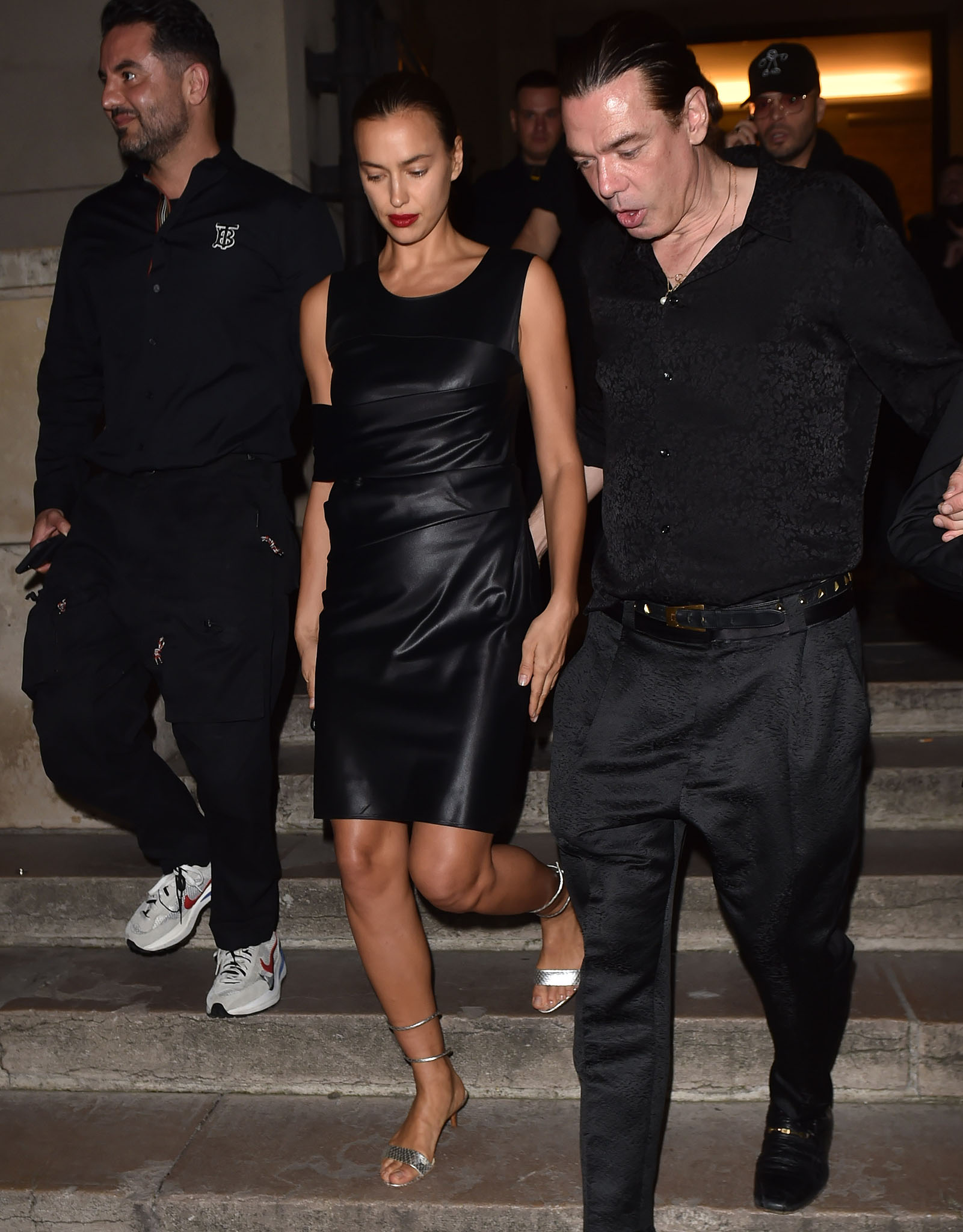 Irina Shayk wearing an archival Helmut Lang Fall/Winter 1997 black leather dress with asymmetric paneling, styled with metallic silver snake-coil sandals featuring wraparound ankle straps and elegant kitten heels