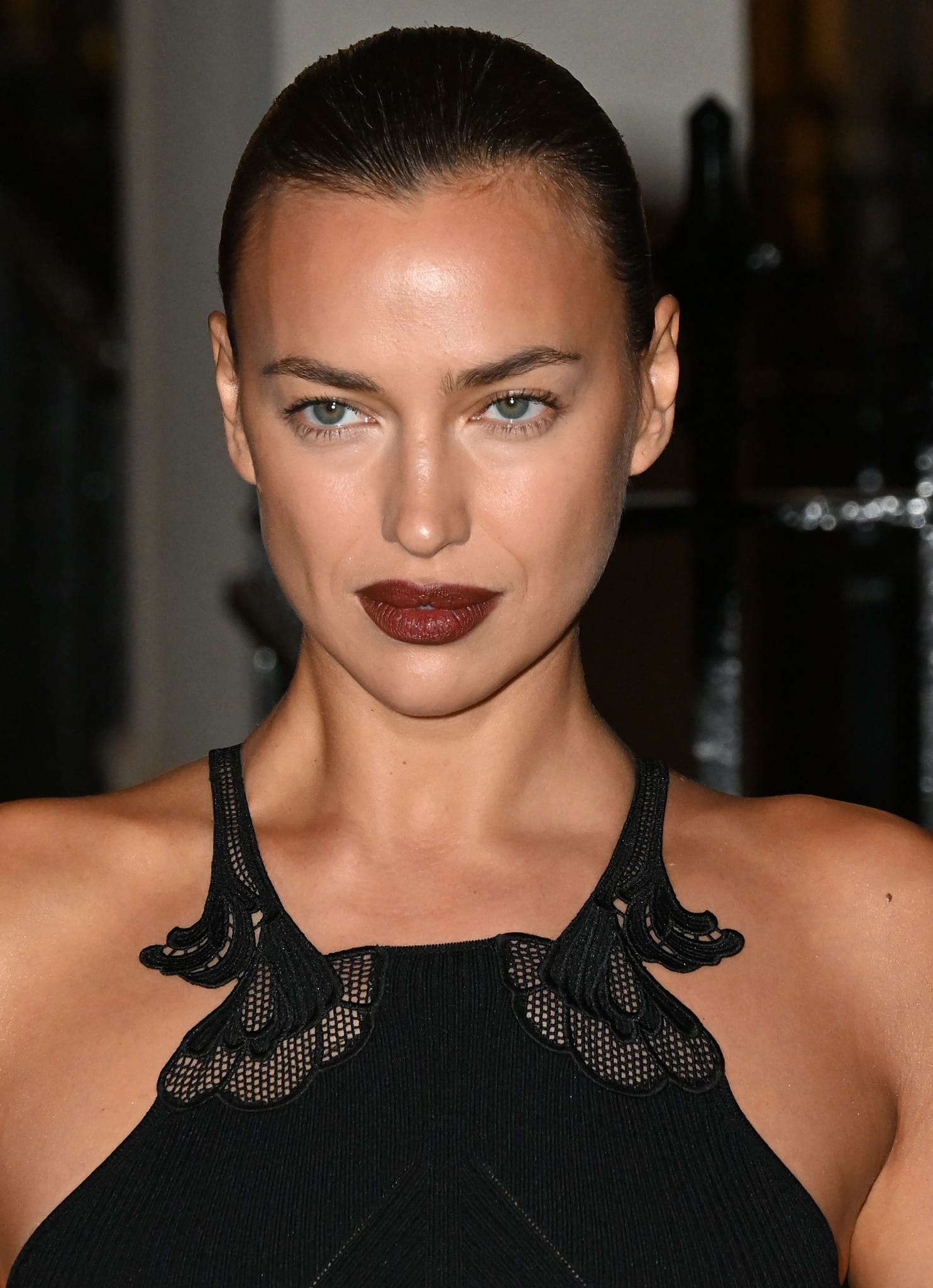 Irina Shayk wears matte brown lipstick with a sleek pulled-back updo, leaving the intricate mesh straps of her black Self-Portrait dress unobstructed