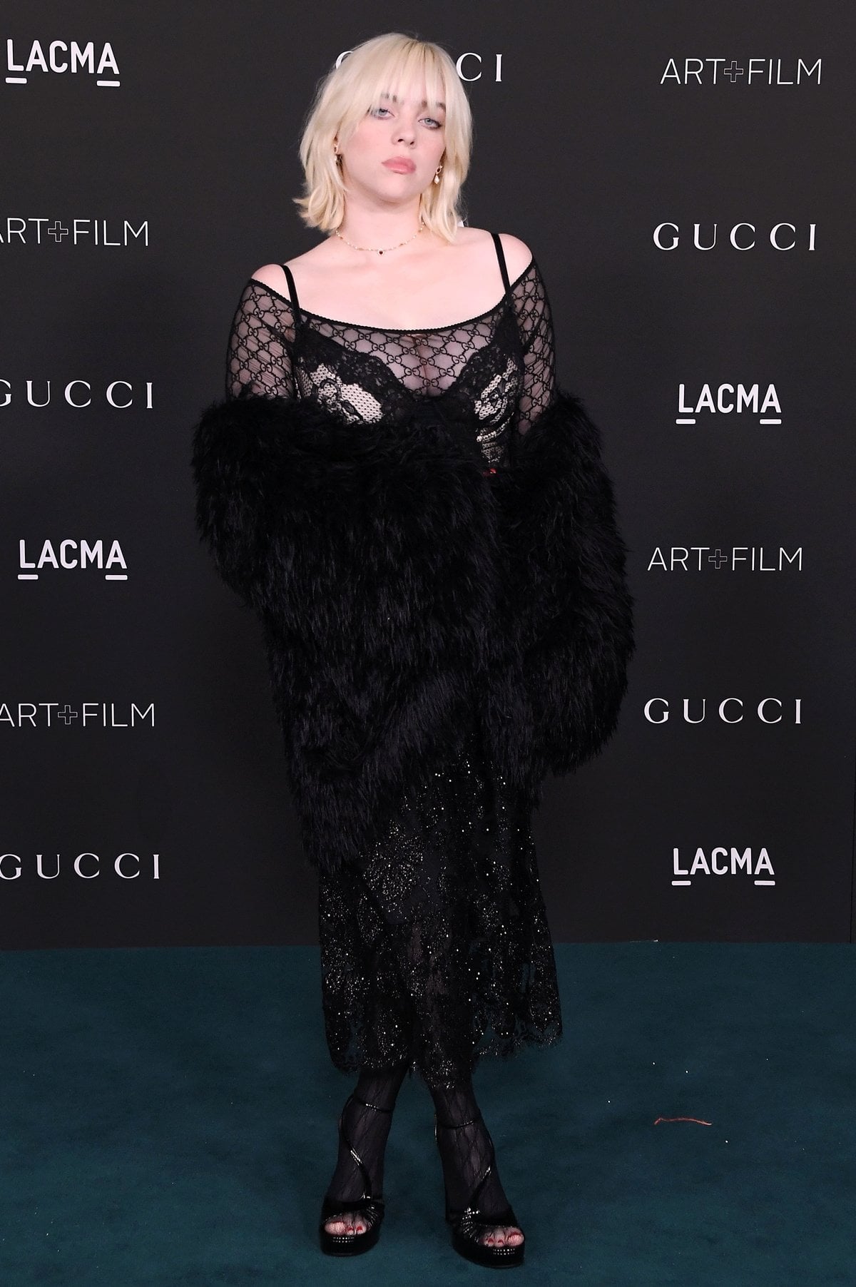 Billie Eilish paired a sheer monogram lace dress and feathered wrap with black open-toe platform sandals and logo stockings that subtly elevate her frame while maintaining a dark, romantic edge
