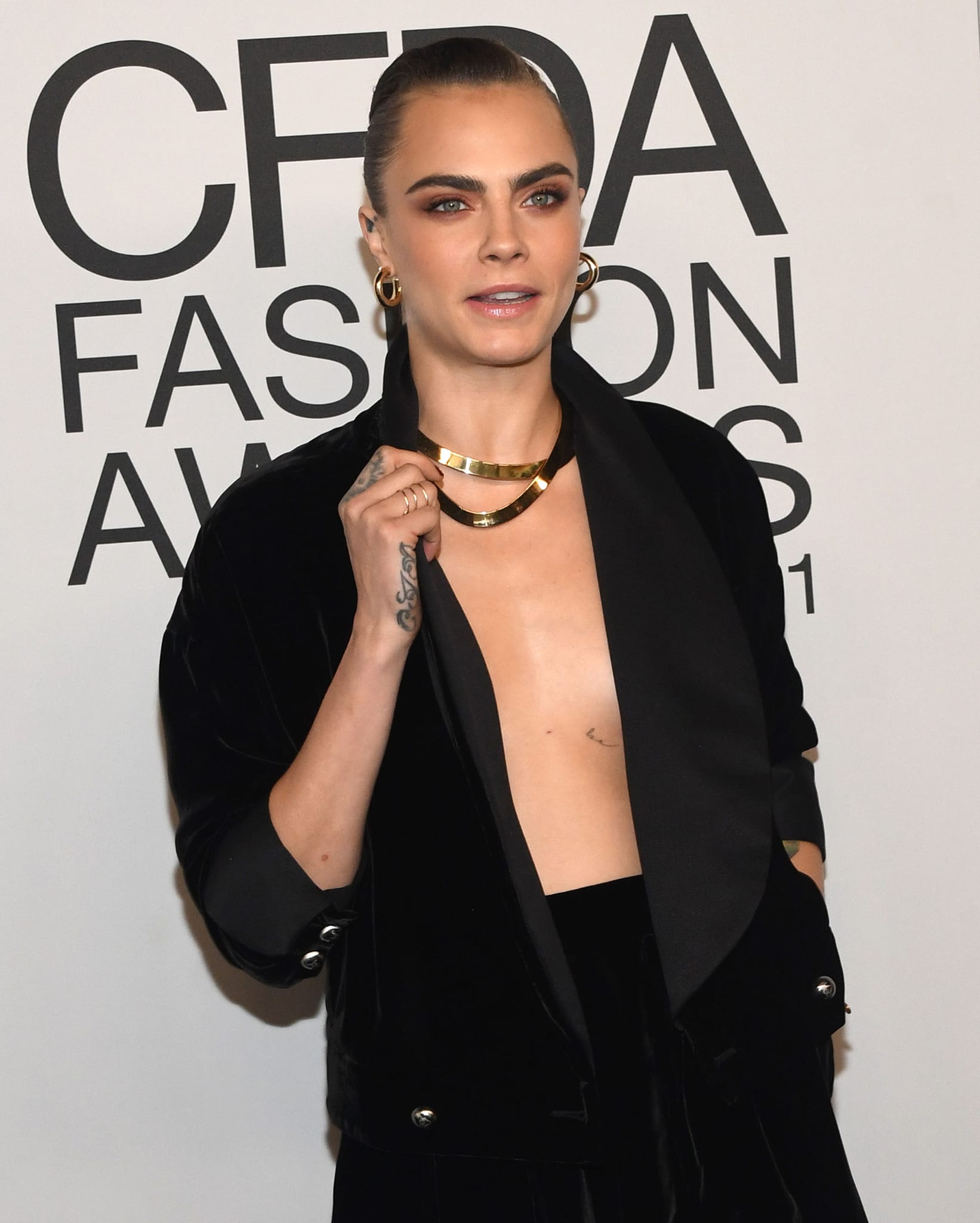 A closer view highlights her sleek bun, smoky eye makeup, and layered gold jewelry, reinforcing the polished edge of the monochrome ensemble
