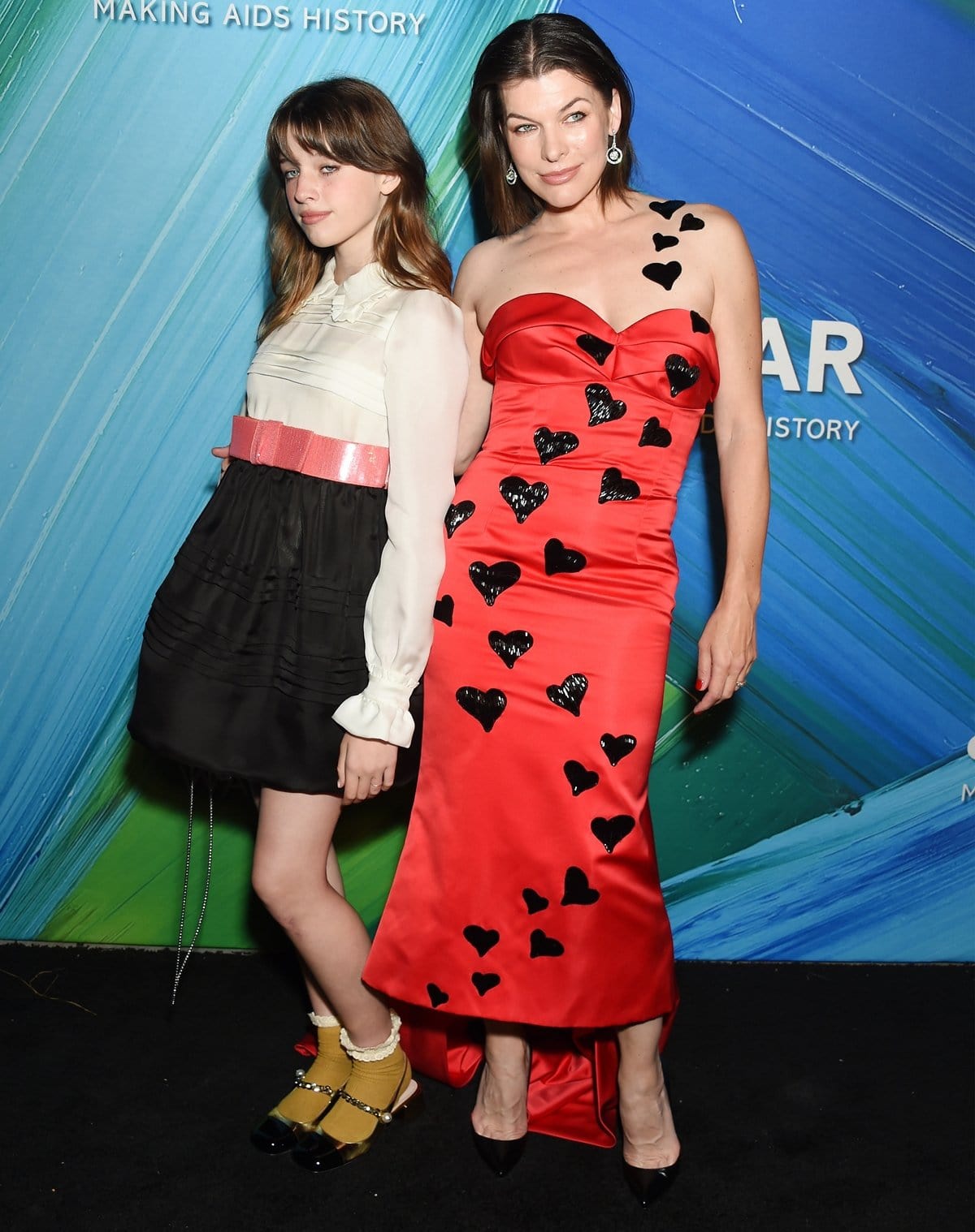 Milla Jovovich and Ever Anderson coordinated in Moschino — Jovovich in a heart-embellished red gown with black pumps and Ever in a ruffled blouse, pink-belted skirt, and chic Mary Jane heels at the 2021 amfAR Gala Los Angeles