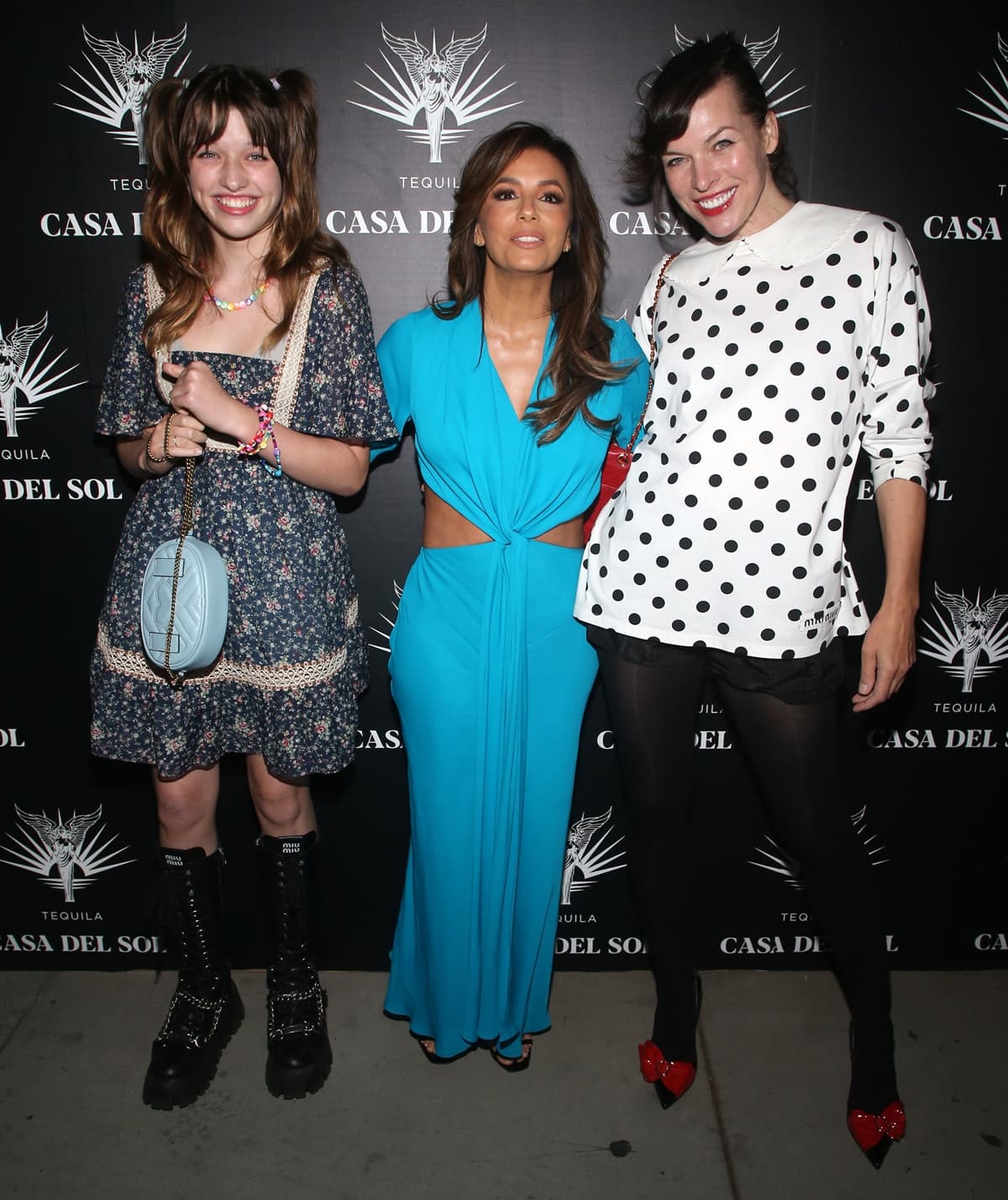 Ever Anderson, Eva Longoria, and Milla Jovovich pose in contrasting styles — Ever in floral combat-boot chic, Longoria in a sleek turquoise gown, and Milla in playful polka dots with red bow heels — at the Drivebys book launch in Los Angeles on October 21, 2021