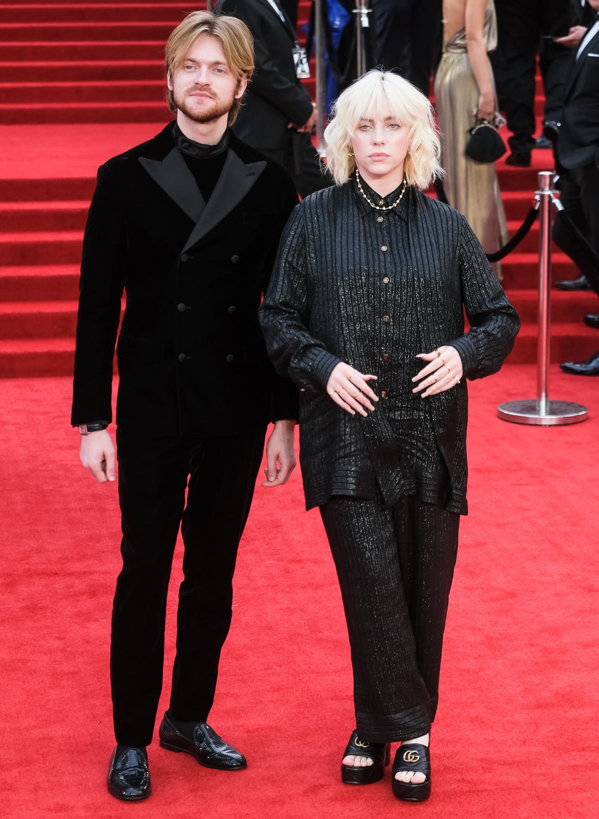 Billie Eilish and Finneas coordinate in black Gucci at the No Time to Die World Premiere on September 28, 2021, at London’s Royal Albert Hall, with Billie grounding her shimmering, oversized shirt-and-trouser set in chunky GG-logo platform slide sandals that add height and a relaxed, modern contrast to the formal red carpet setting