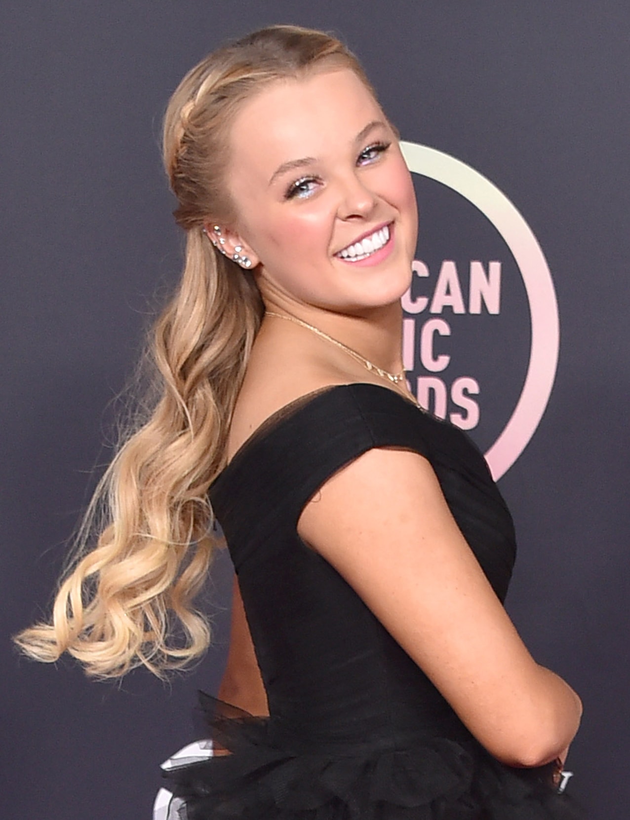 JoJo Siwa flashes a radiant smile over her shoulder in a black off-the-shoulder gown, styling her sleek half-up waves with sparkling ear cuffs and delicate gold jewelry for a polished red-carpet moment
