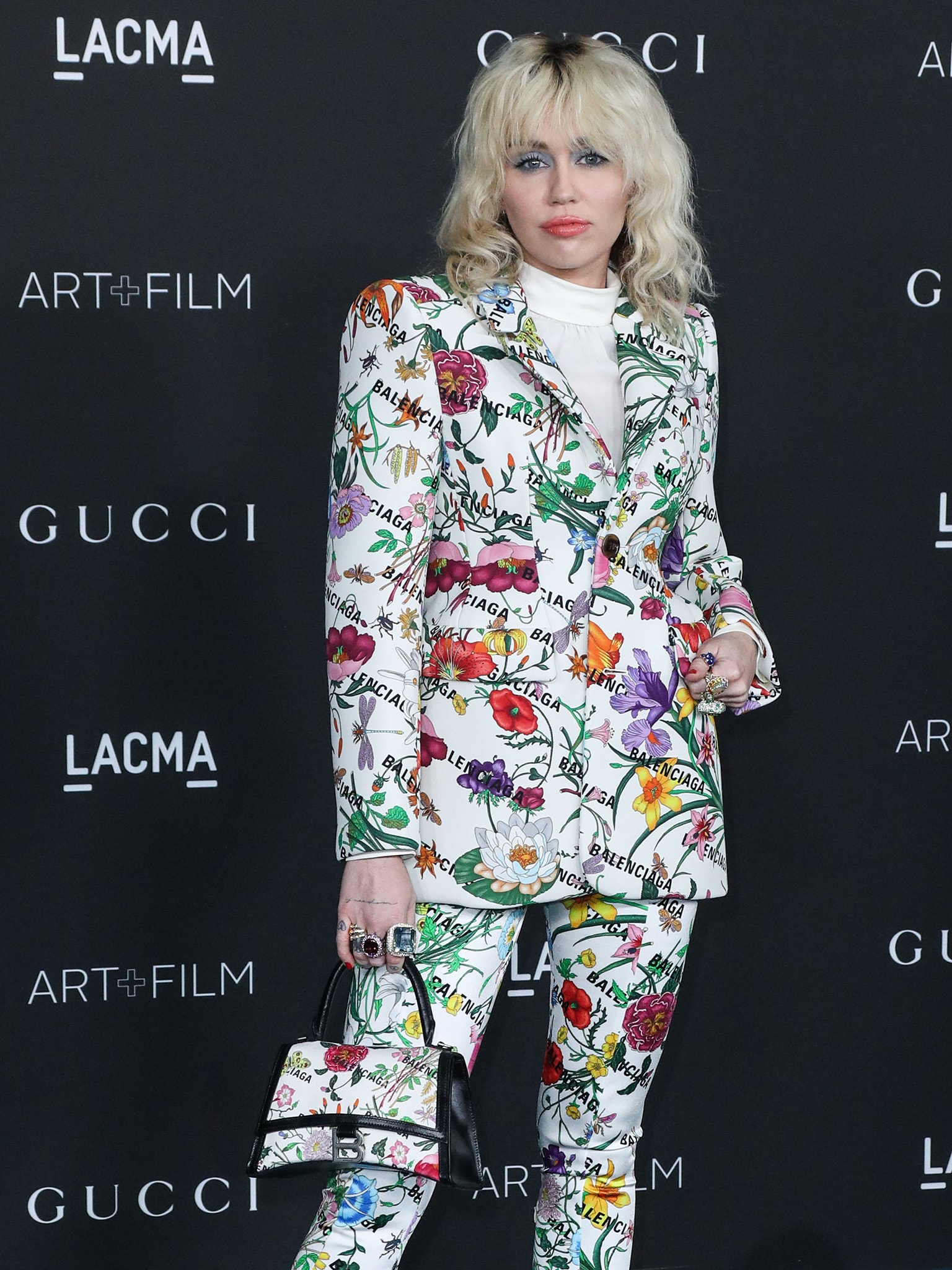 Miley Cyrus complements her Gucci x Balenciaga Hacker Project pantashoes with a matching floral handbag and oversized gemstone rings, reinforcing the collection's head-to-toe maximalist aesthetic