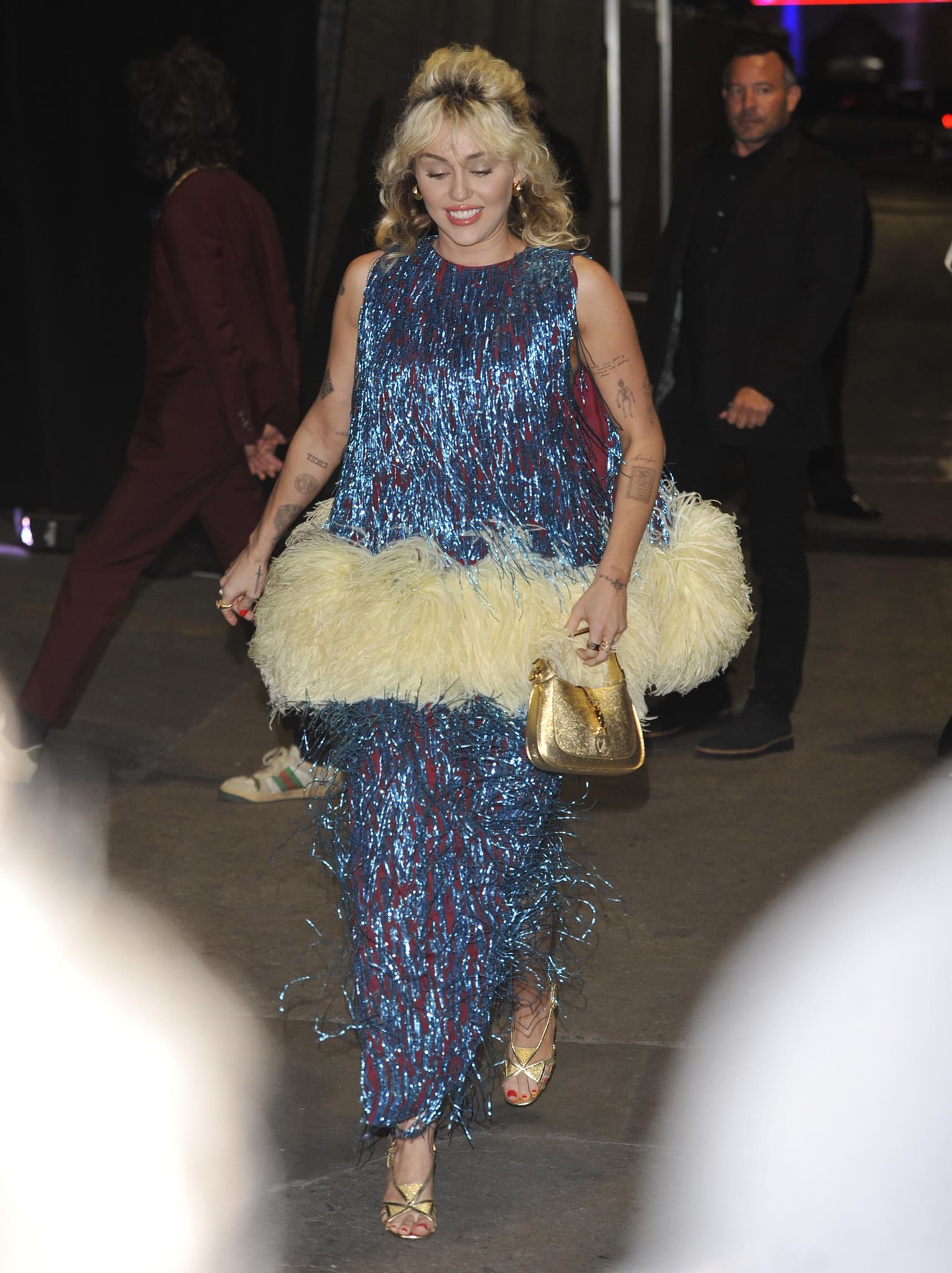 Miley Cyrus arrives at Gucci's Love Parade fashion show in Hollywood on November 2, 2021, wearing a shimmering blue fringe dress with oversized feather trim, metallic gold Gucci sandals, and a matching Mini Jackie bag