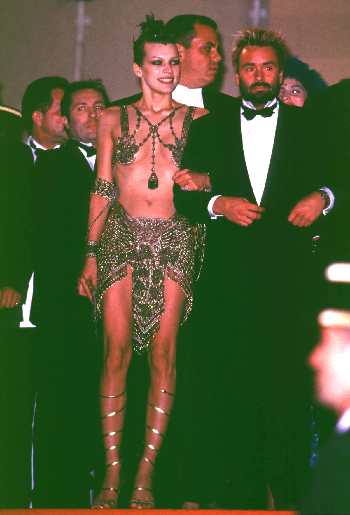 Milla Jovovich stuns in John Galliano’s metallic chainmail design from Dior’s Fall 1997 collection, paired with silver gladiator heels, at the Cannes Film Festival alongside director Luc Besson