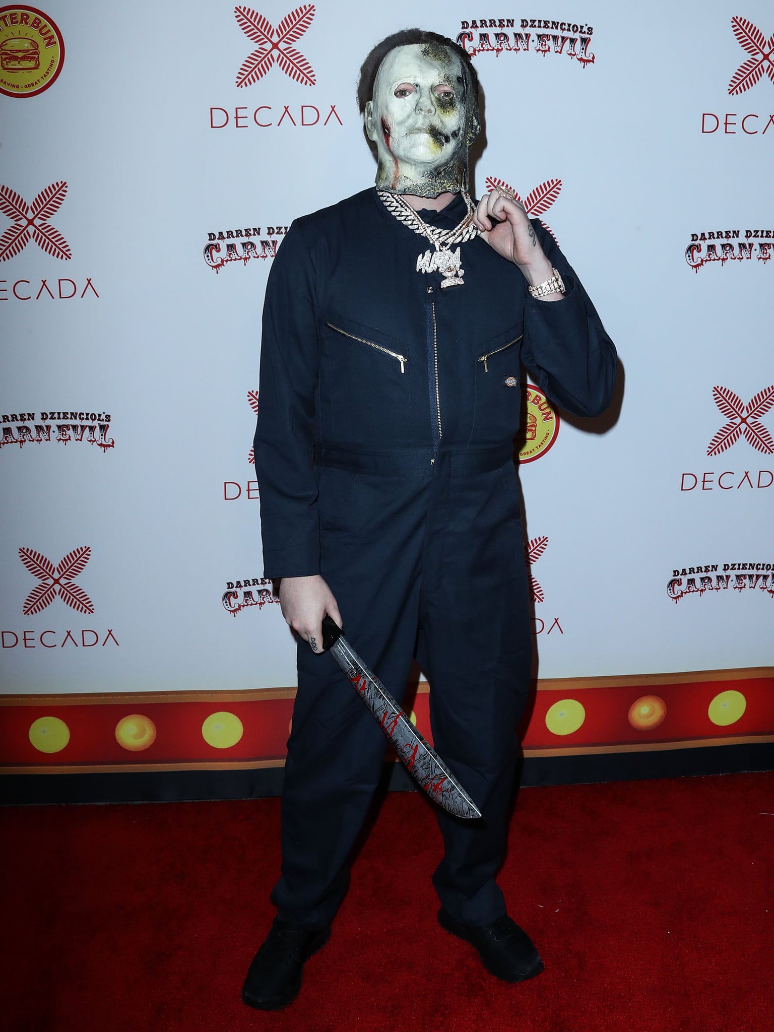 Murda Beatz embraces slasher-movie menace as Michael Myers in a navy zip-front jumpsuit and eerie mask, grounding the chilling look with understated black lace-up shoes that keep the focus on the haunting costume and prop blade