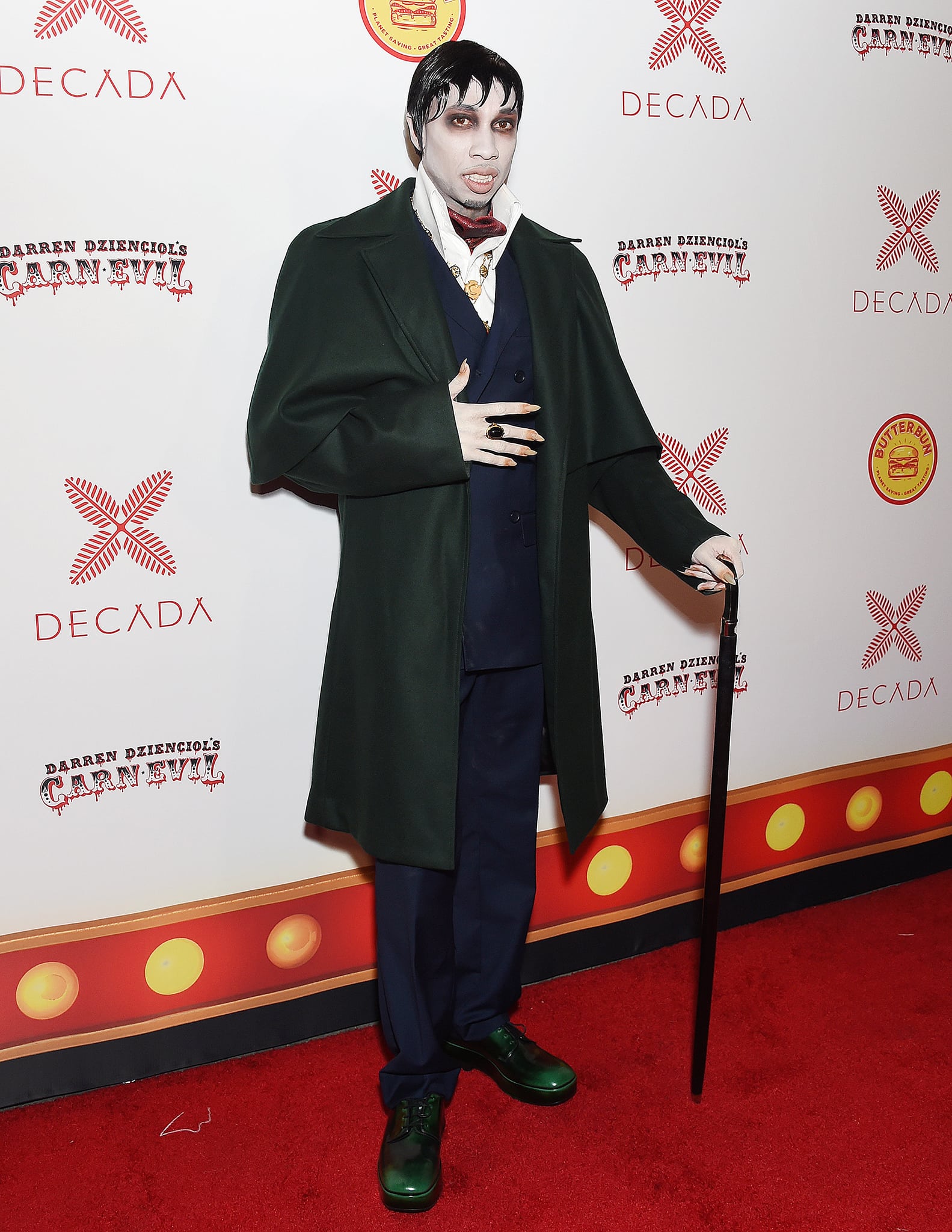 Tyga channels gothic drama as Barnabas Collins in a dark three-piece suit layered under a sweeping green overcoat, completing the vintage vampire look with polished green leather dress shoes and a classic black cane for theatrical flair
