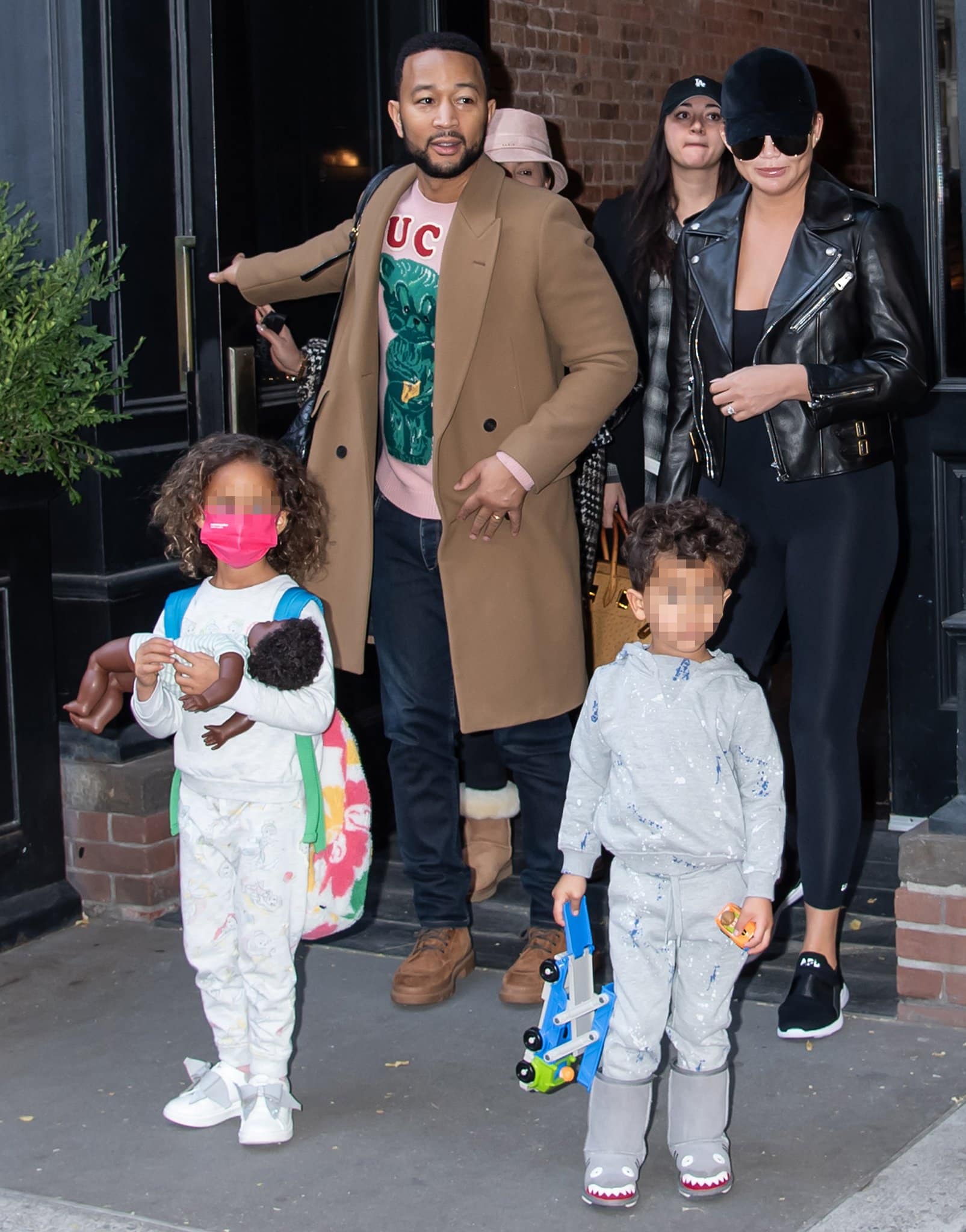 John Legend layers a camel overcoat over a graphic sweater and tan suede boots, while Chrissy Teigen keeps it sleek in black athleisure with slip-on APL “Techloom Bliss” sneakers as they head to a New York City airport with Luna and Miles on December 5, 2021