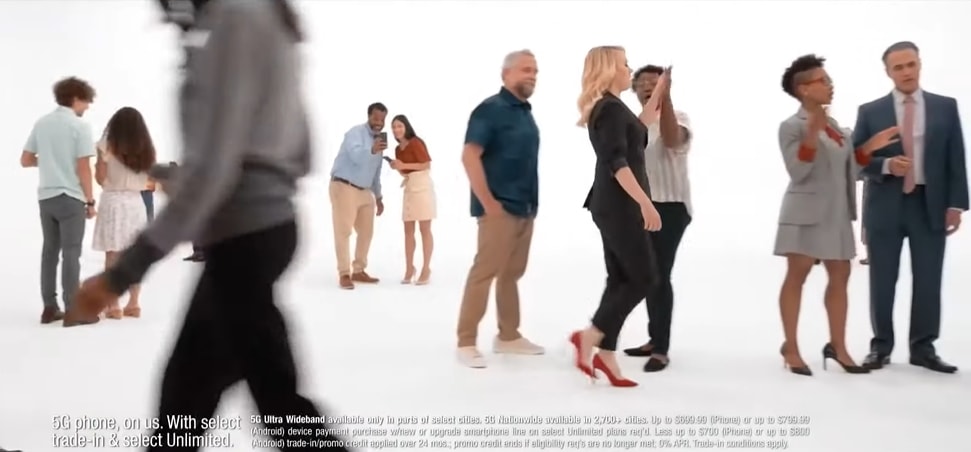 Kate McKinnon struts through a minimalist Verizon set in sleek red pumps and a tailored black jumpsuit — a tongue-in-cheek nod to her off-balance comedic charm and confident heel game