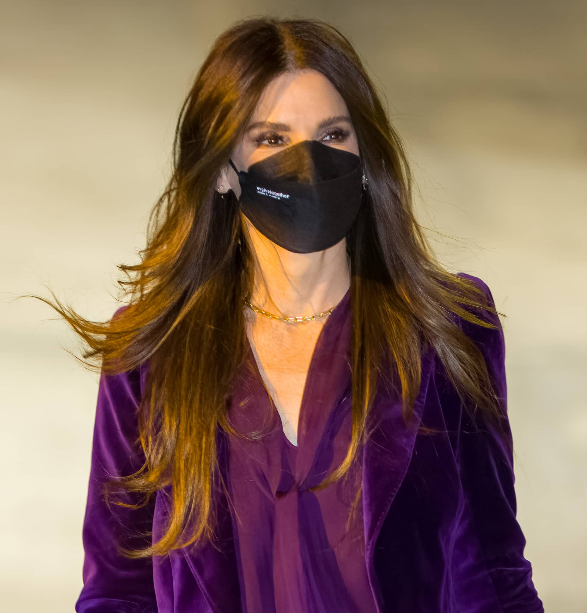 Bullock complements the rich plum velvet with center-parted caramel-toned waves, neutral smoky eye makeup, and a delicate gold chain necklace