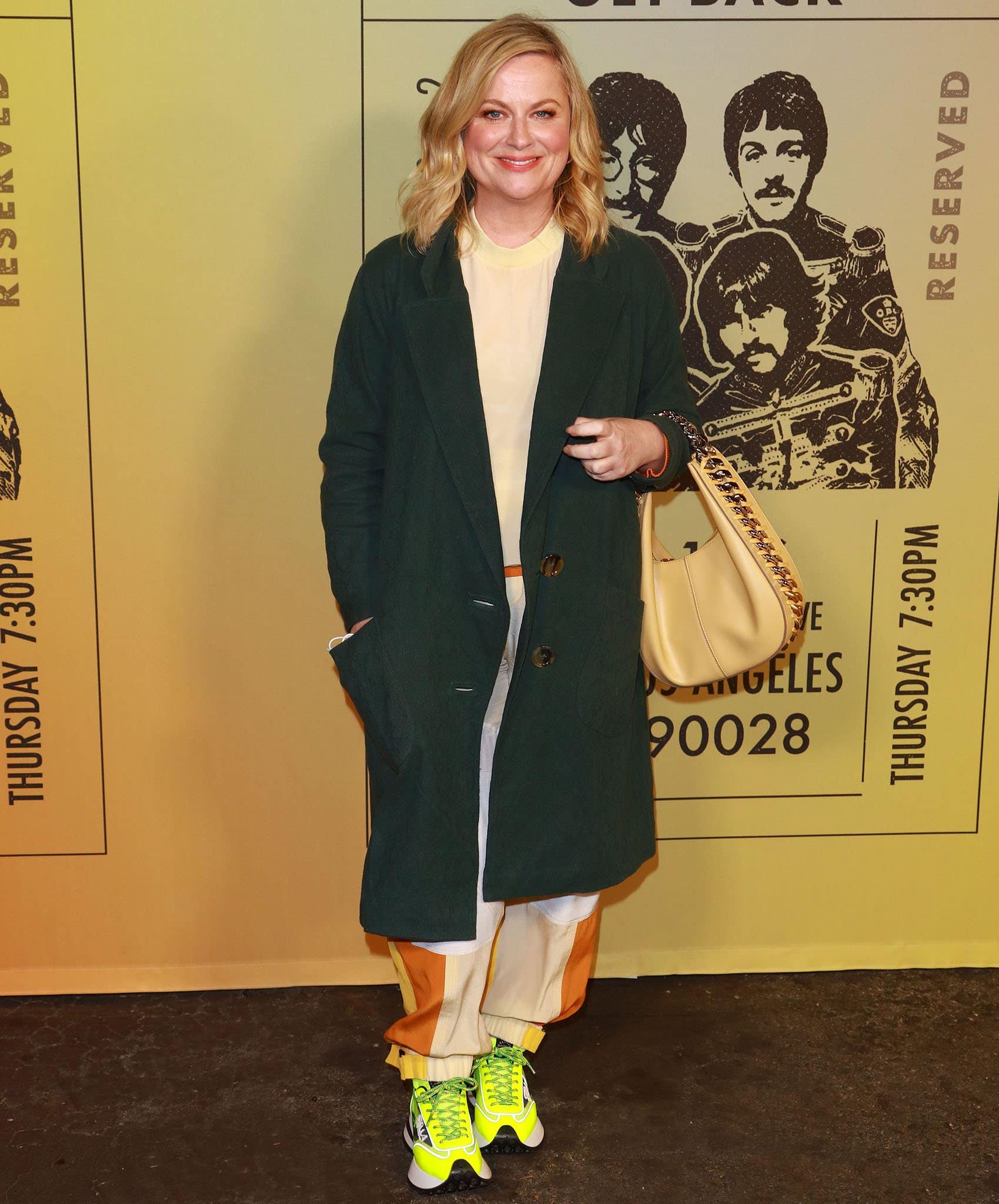 Amy Poehler keeps it cool and casual in a forest green coat layered over a neutral top and color-block joggers, finishing the look with eye-catching neon yellow sneakers that add a playful pop of energy