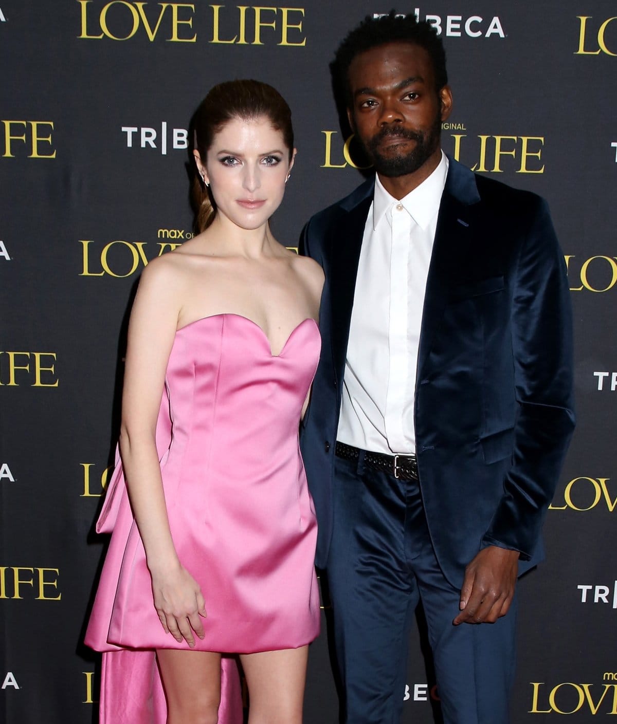 Anna Kendrick pairs her satin pink mini dress with pointed-toe pumps, using a high hemline and sharp toe shape to elongate her petite 5'1" frame beside taller co-star William Jackson Harper