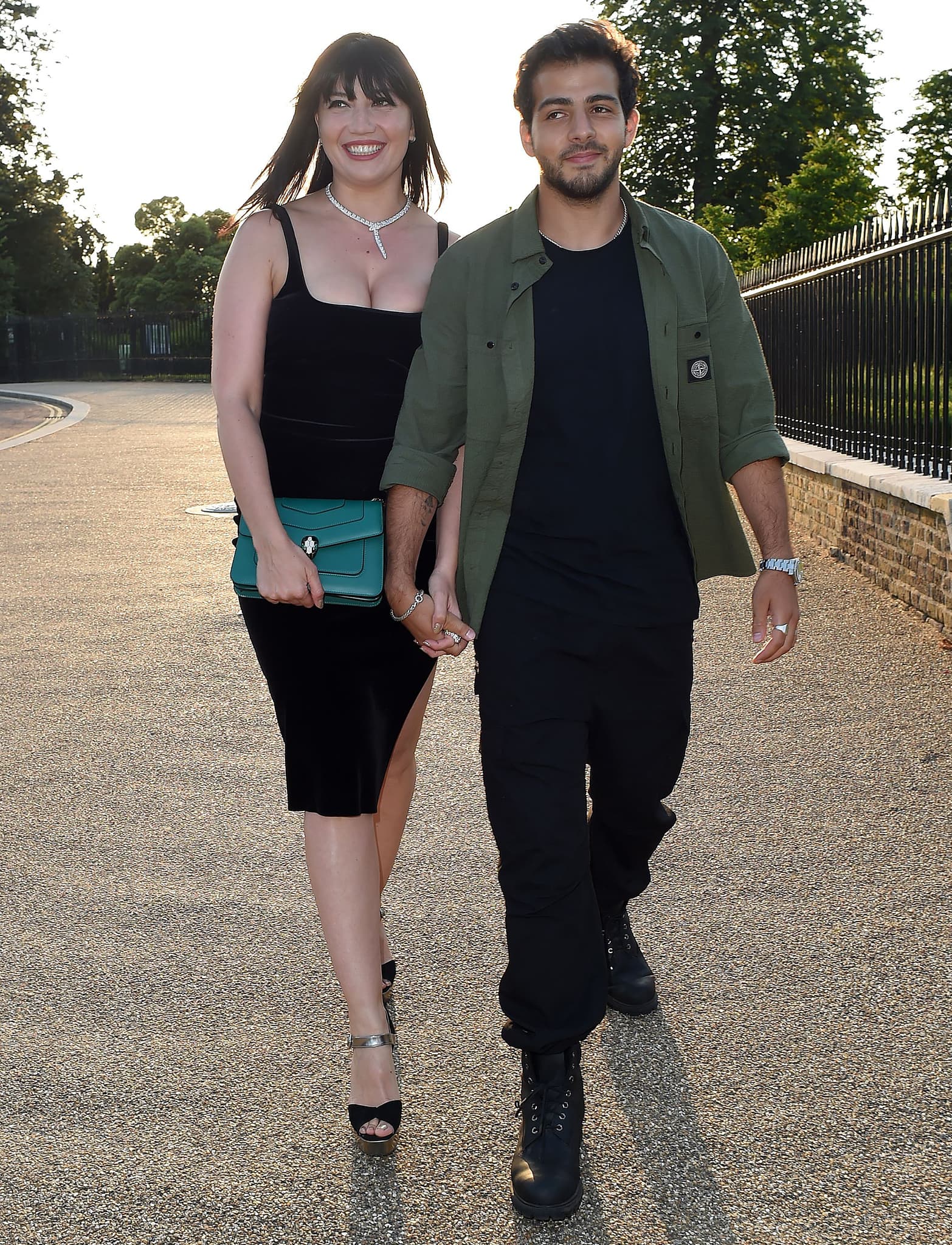 Daisy Lowe arrives at the Bulgari Summer Party in Hyde Park on July 22, 2021, wearing a sleek black midi dress with a thigh-high slit, paired with metallic ankle-strap platform sandals that add height and edge, while Jordan Saul keeps it relaxed in black boots and a utility-inspired look
