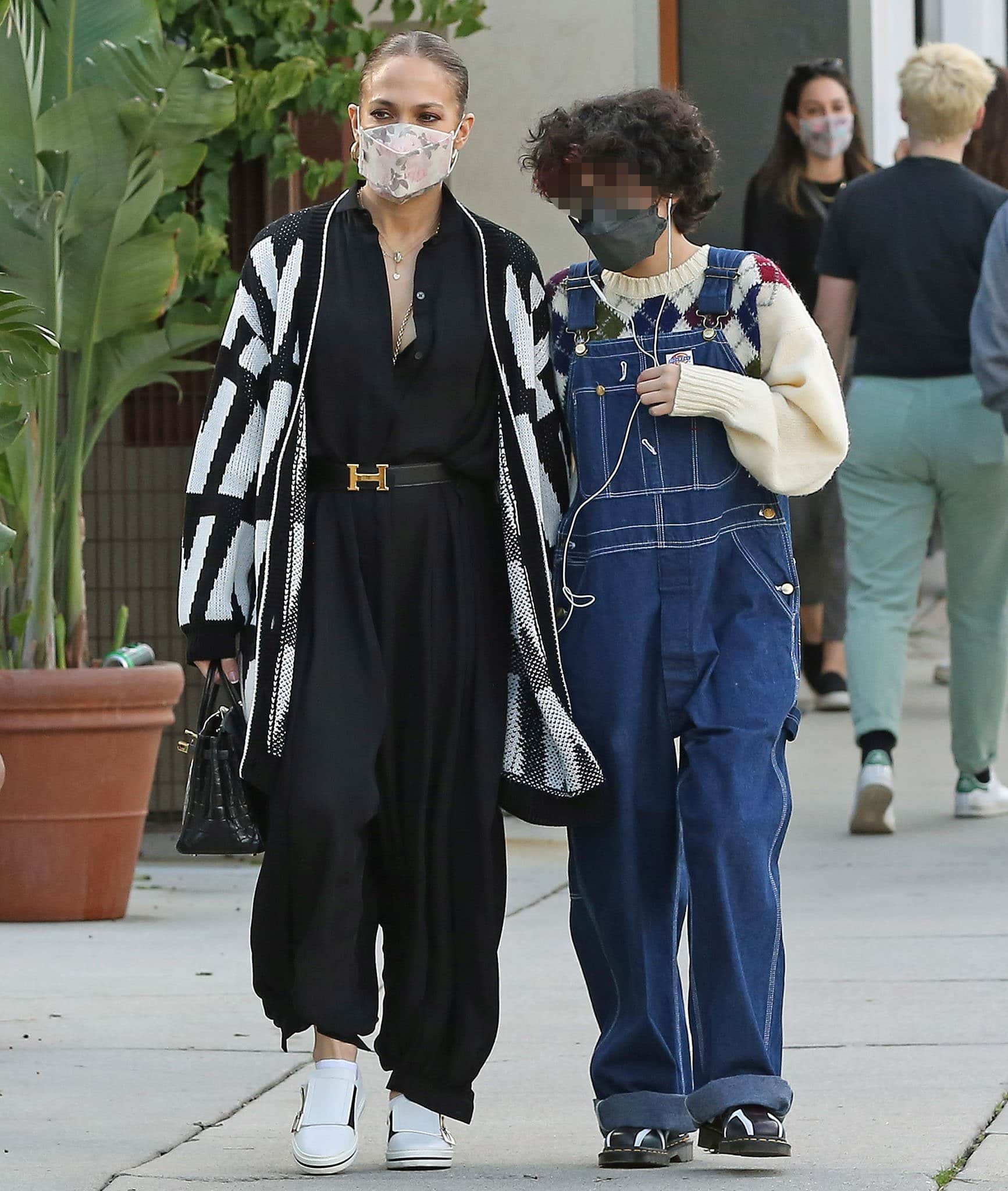 Jennifer Lopez balances flowing proportions with structured accessories in a relaxed black jumpsuit layered under a bold Valentino cardigan, finishing the monochrome look with crisp white slip-on shoes and an Hermès belt