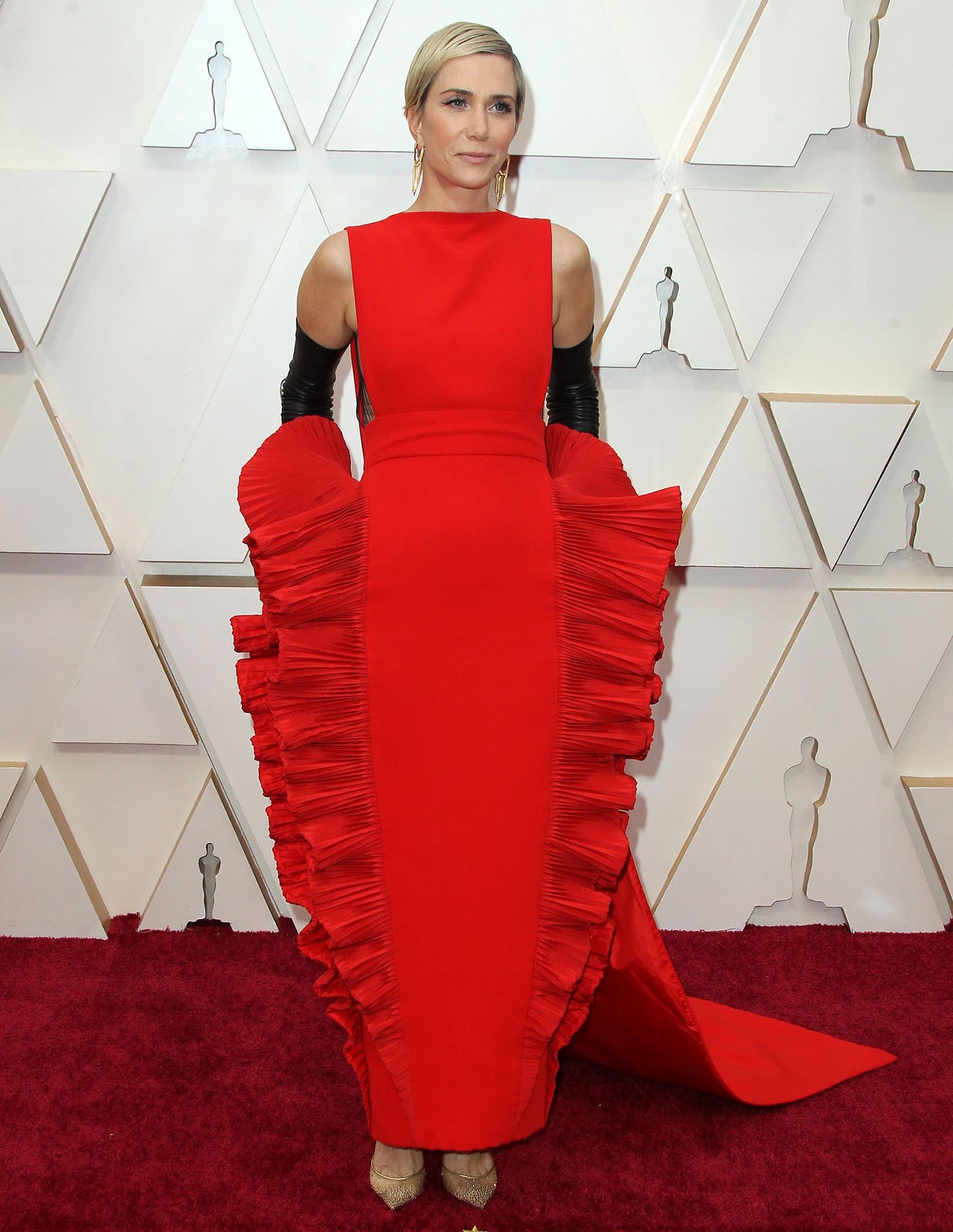 Kristen Wiig commands attention on the Oscars red carpet in a sculptural red Valentino gown with dramatic pleated ruffles, styled with opera-length black gloves and gold pointed-toe pumps for a daring high-fashion moment