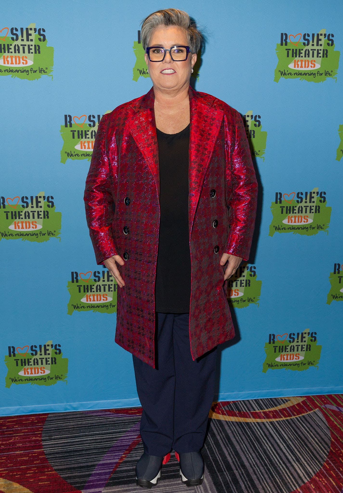 Rosie O'Donnell adds flair to her signature style with a shimmering red jacquard coat, pairing it with relaxed navy trousers and unique split-toe sneakers at a Rosie’s Theater Kids event