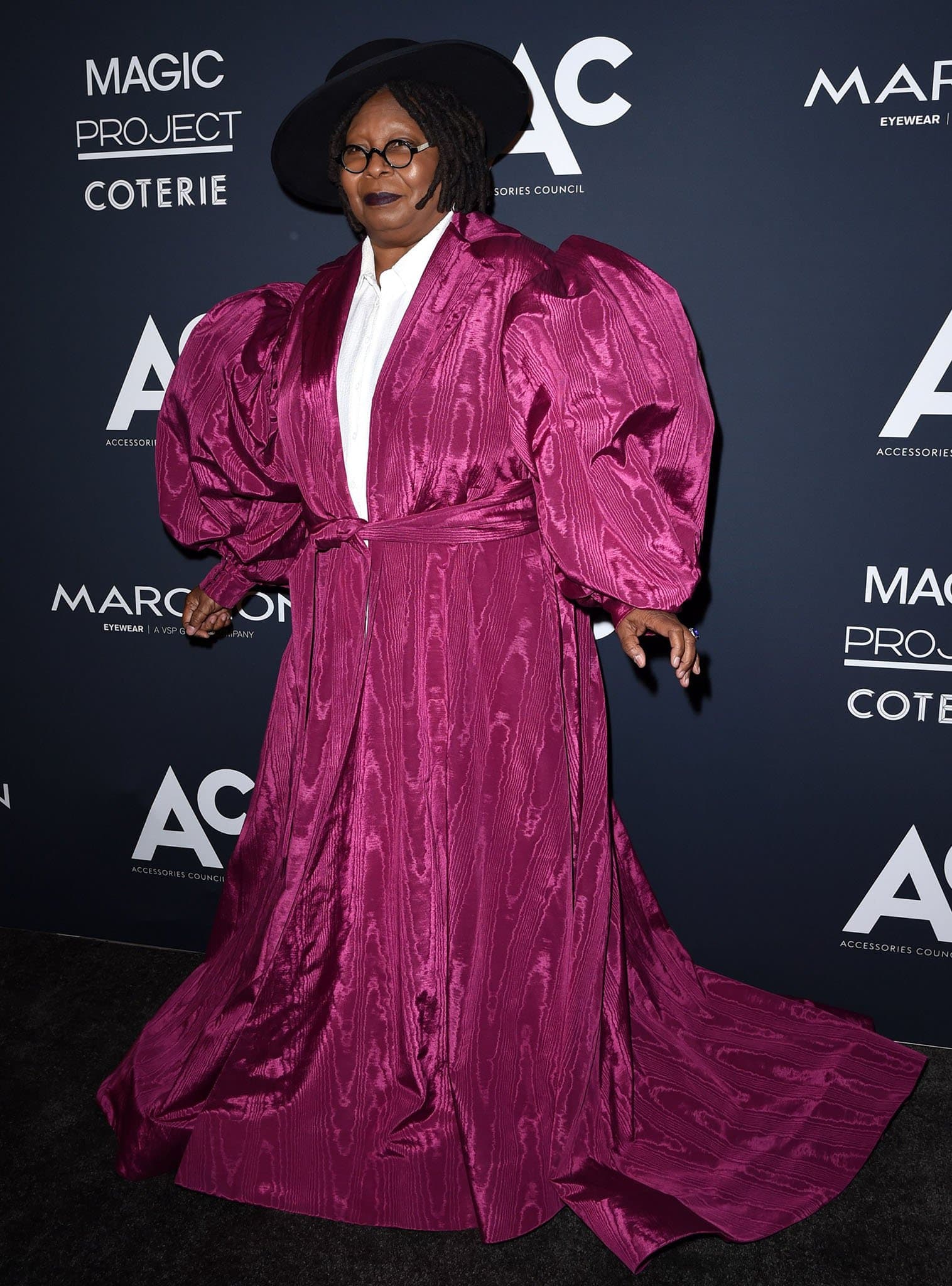 Whoopi Goldberg makes a dramatic entrance in a voluminous fuchsia moiré Christian Siriano gown with exaggerated puff sleeves, paired with a wide-brimmed black hat and round glasses for a bold, fashion-forward statement at the Accessories Council event