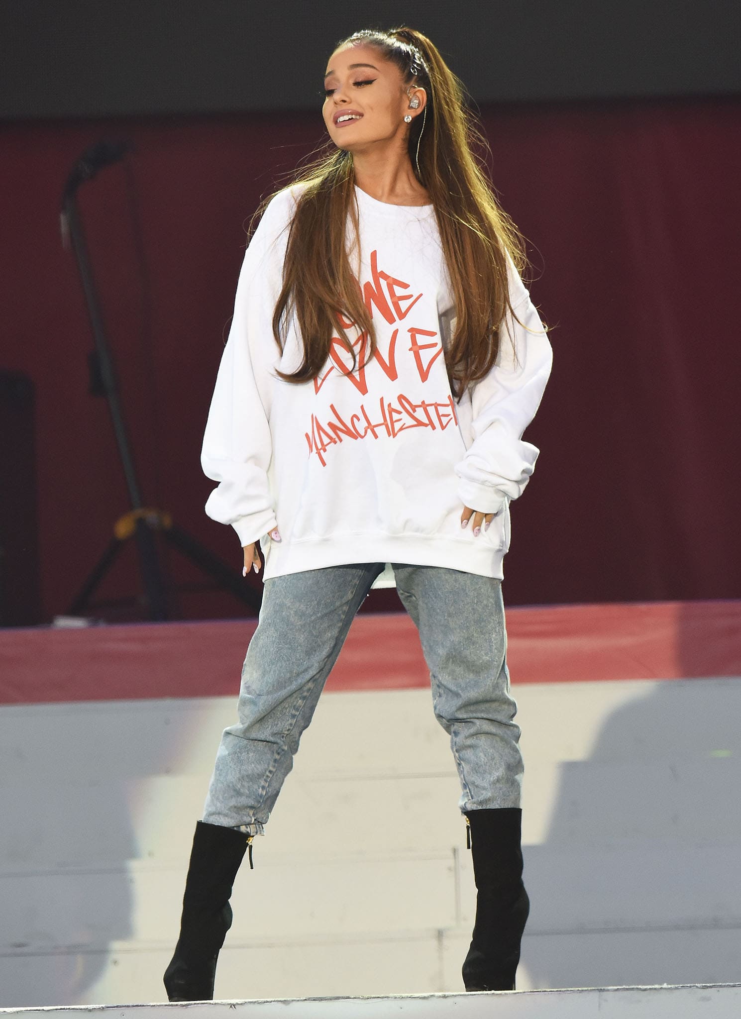 Ariana Grande performs at the One Love Manchester benefit concert in an oversized “One Love Manchester” sweatshirt, relaxed jeans, and black heeled boots