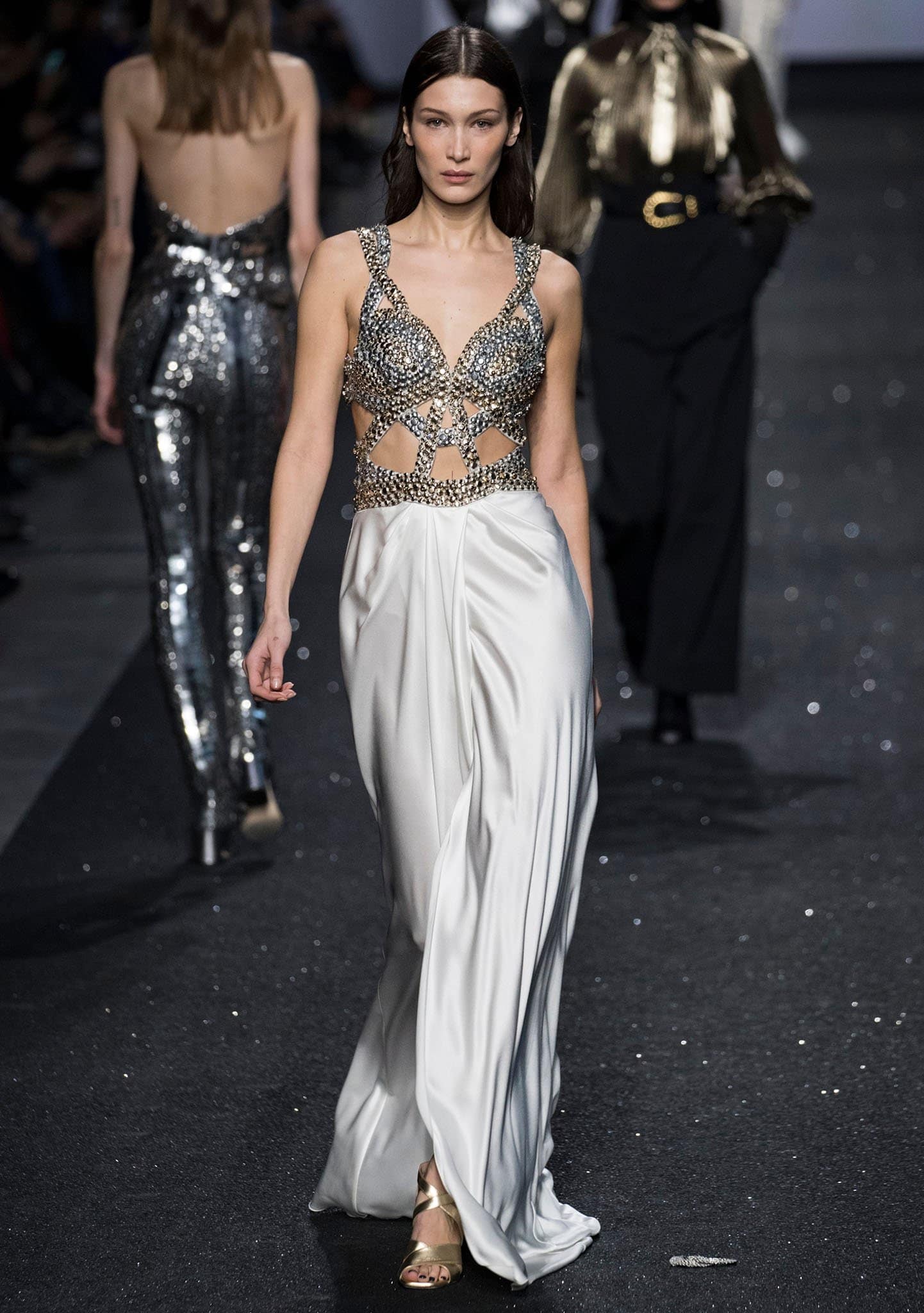 Standing at 5 feet 8 inches (172.7 cm), Bella Hadid captivates in a crystal-embellished cutout bodice and flowing white satin skirt, elevated by metallic gold peep-toe heels that add height and glamour to her runway presence