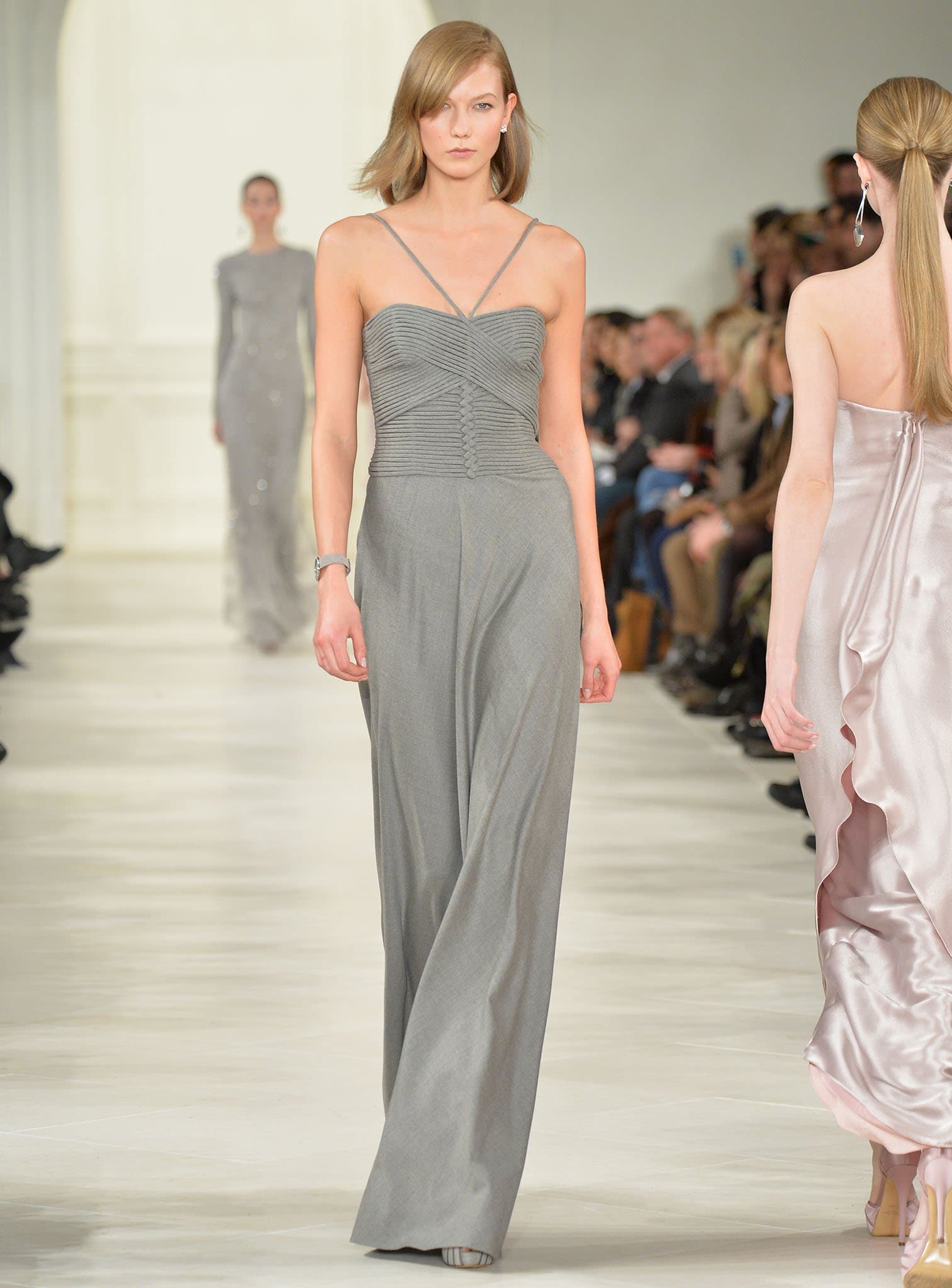 Karlie Kloss, standing tall at 6 feet 1 ½ inches (186.7 cm), glides down the runway in a sophisticated gray strappy jumpsuit with flowing wide-leg pants, her statuesque frame and poised walk embodying modern elegance