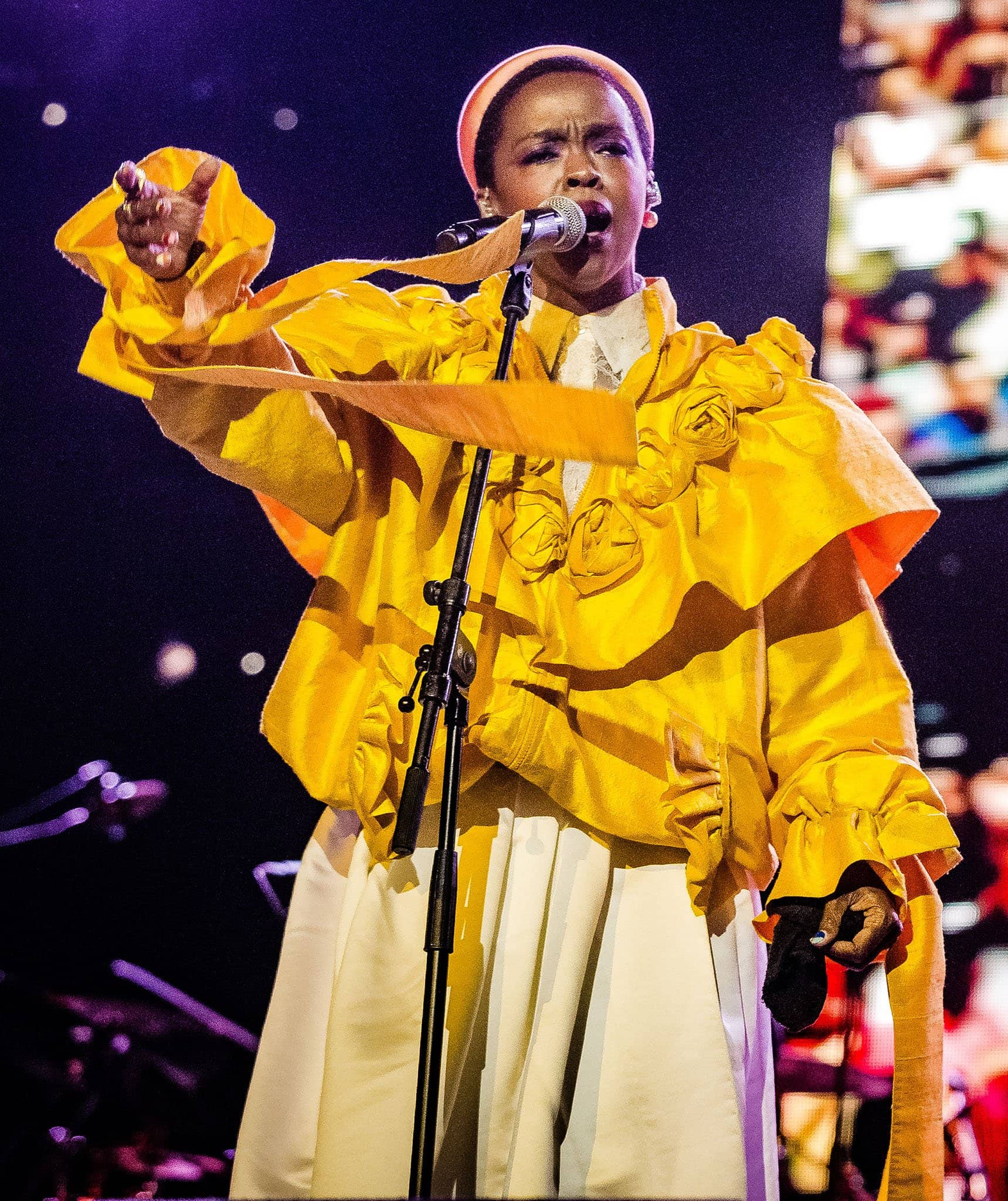 Lauryn Hill commands the stage in a dramatic yellow ruffled jacket adorned with rosette detailing, paired with a voluminous white skirt and matching headband for an artful, statement-making performance look