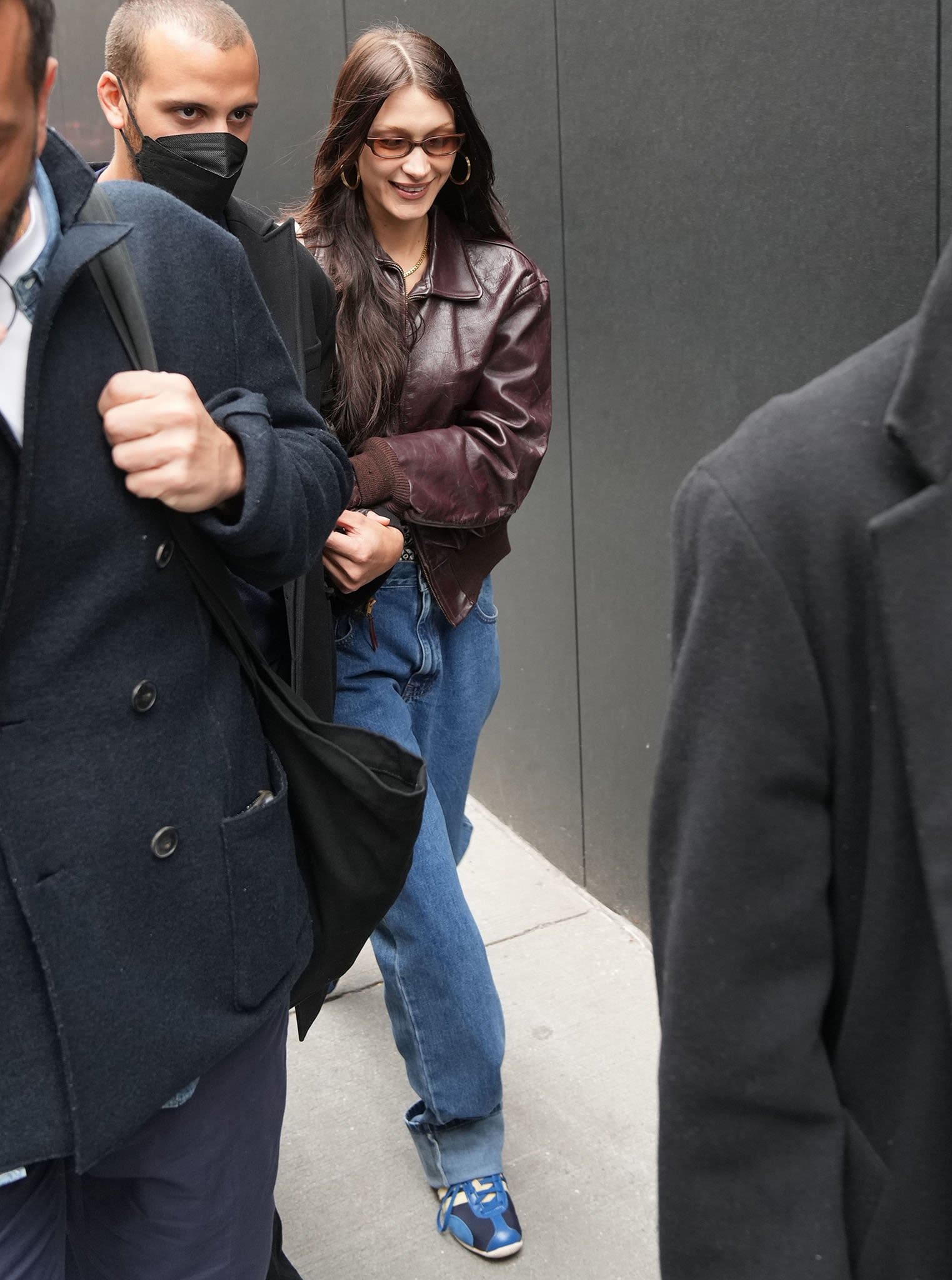 Bella Hadid steps out in a rich maroon leather biker jacket and relaxed blue jeans, grounding the off-duty look with retro-inspired blue sneakers that add a sporty, early-2000s edge