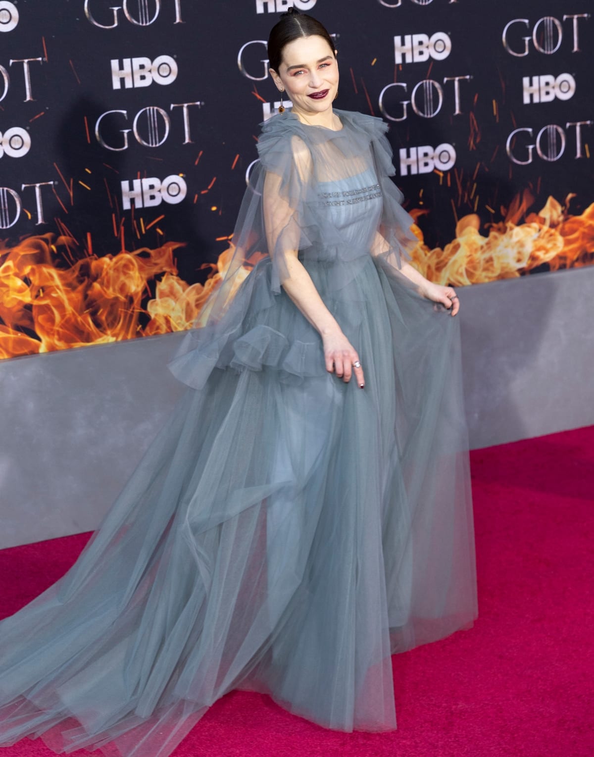 Emilia Clarke softens the drama of her ethereal teal-blue Valentino tulle gown with minimal, likely nude-toned sandals that visually disappear under the sheer layers—an intentional styling move that lengthens the leg line and keeps the focus on the gown’s weightless volume and gemstone-embellished bodice at the Game of Thrones Season 8 premiere in New York City on April 3, 2019