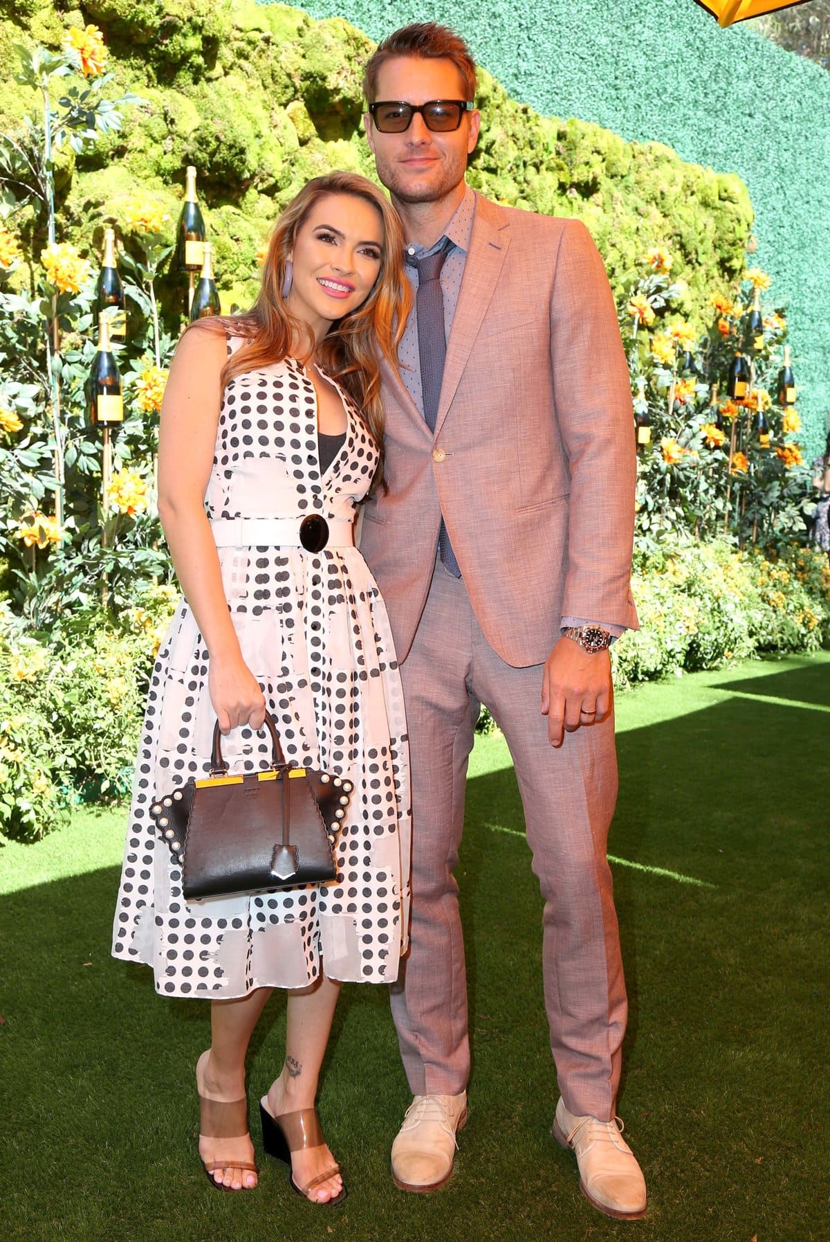 Justin Hartley and Chrishell Stause pose together at a garden event, with Stause wearing a black-and-white polka-dot midi dress cinched with a statement belt and paired with transparent strap sandals, while Hartley keeps it polished in a dusty-rose suit and suede lace-up shoes