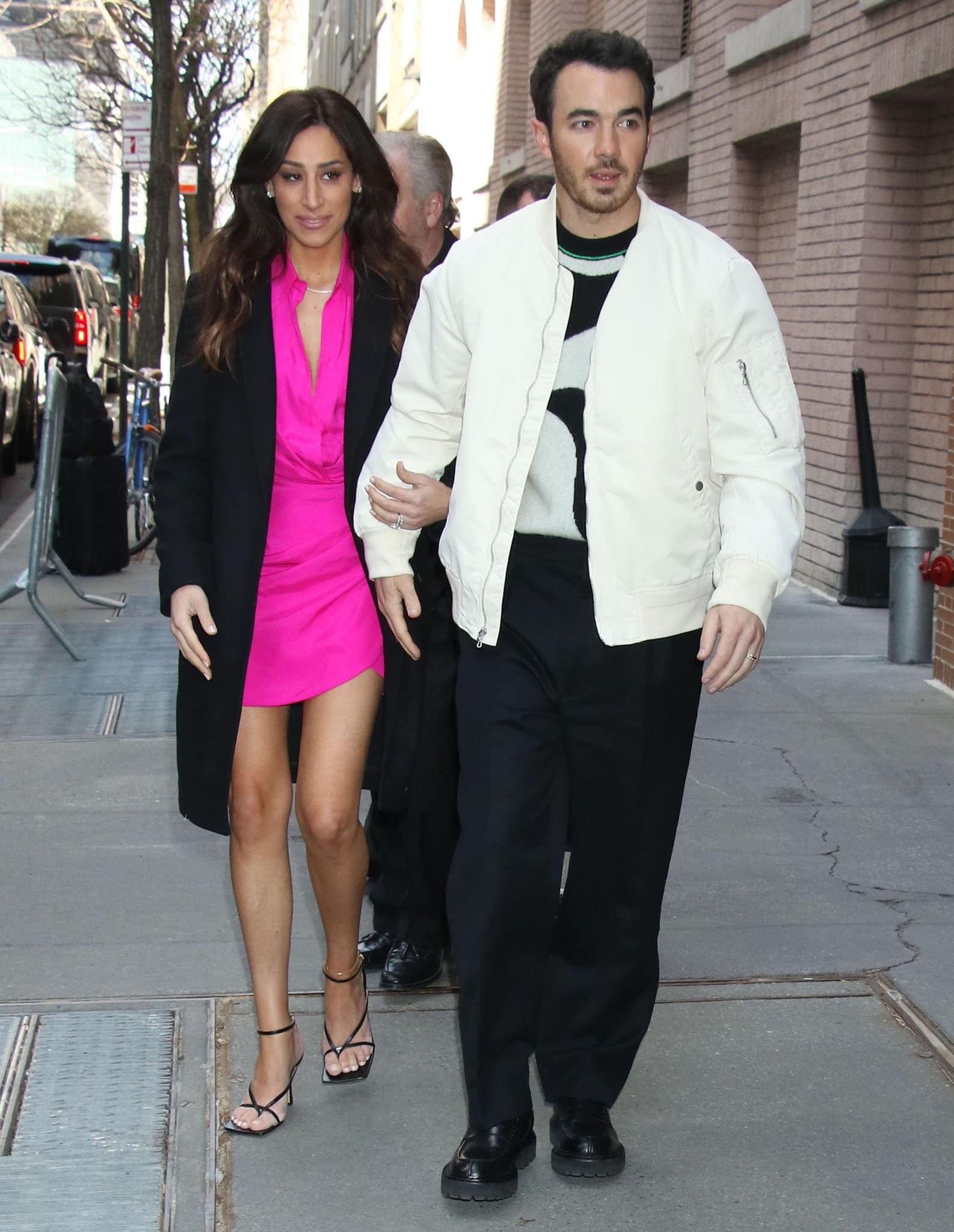 Danielle Jonas layers a black coat over a vibrant pink mini dress, finishing the look with black Bottega Veneta Stretch sandals as she arrives at The View with Kevin Jonas during their book promotion tour