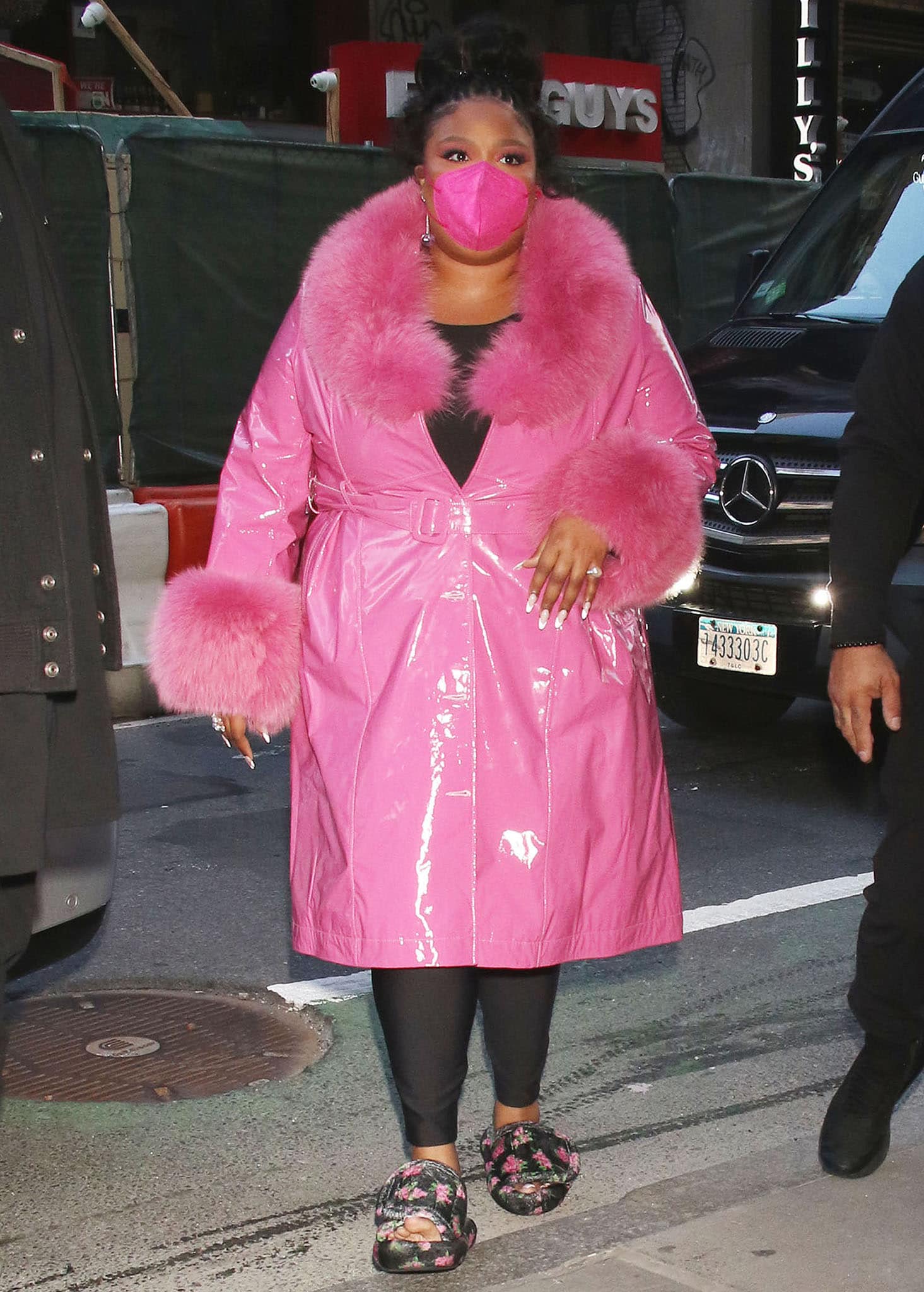 Lizzo wears a glossy pink faux-fur-trimmed coat over a black Yitty bodysuit and leggings with floral slide sandals outside The Today Show on April 11, 2022