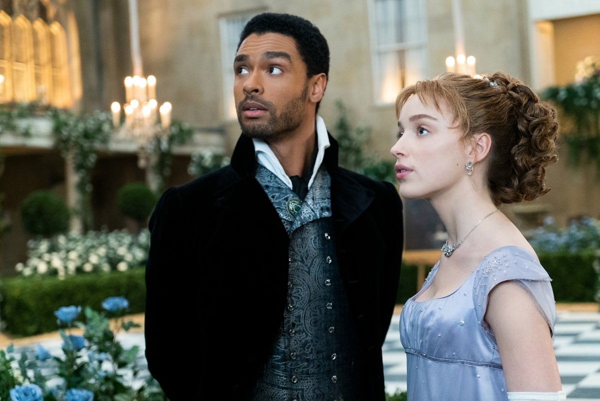 Bridgerton stars Phoebe Dynevor and Regé-Jean Page share a scene as Daphne Bridgerton and Simon Basset, dressed in opulent Regency-era attire amid a lavish garden party setting