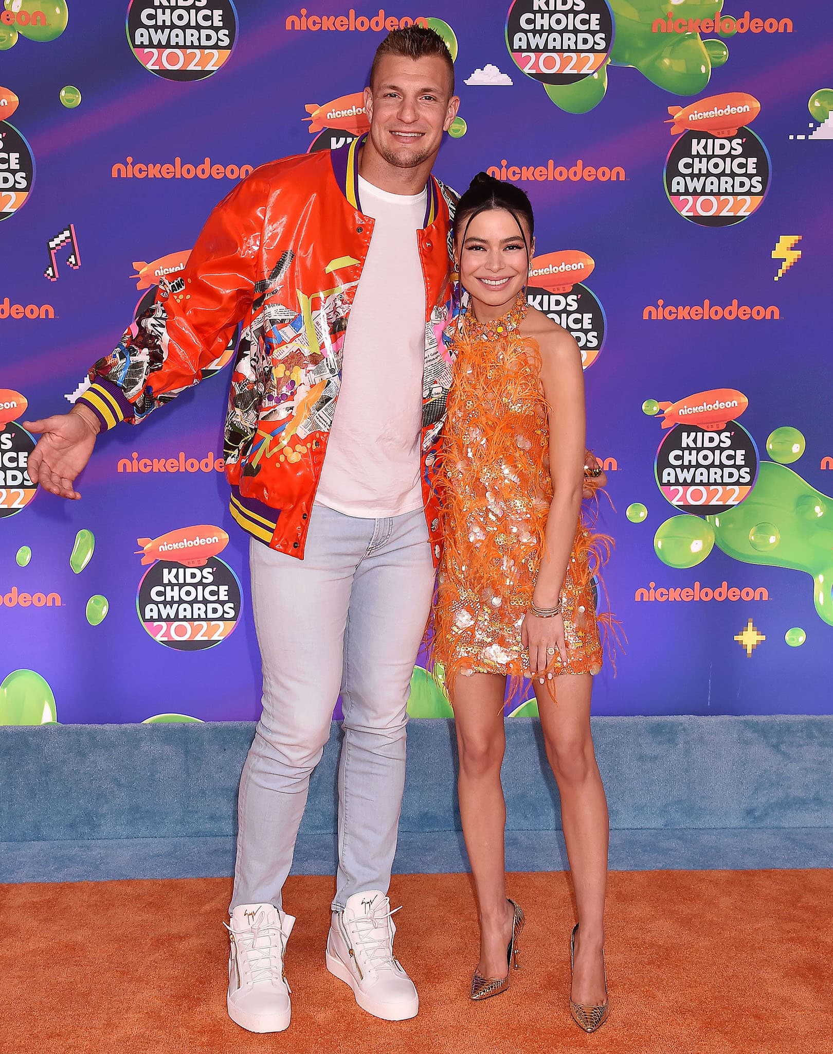 Rob Gronkowski, who stands at an imposing 6′6¼″ (1.99 m), towers over Miranda Cosgrove as the 2022 Kids’ Choice Awards co-hosts pose on the orange carpet, her sculptural gold Voyette Lee pumps subtly boosting her petite frame beside the former NFL star