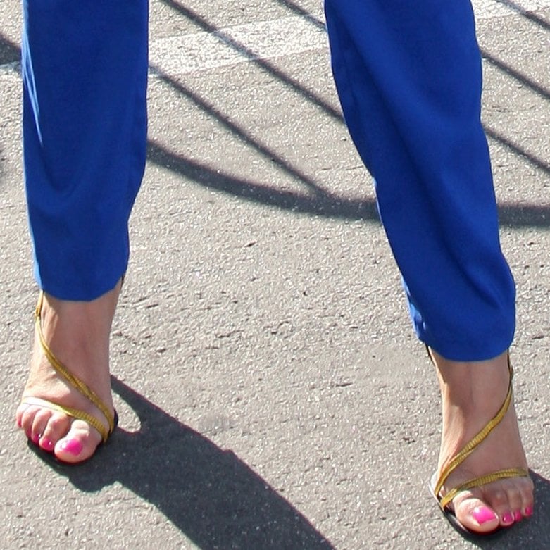 The image shows a close-up of feet in strappy sandals, revealing noticeable bunions and bright pink-painted toenails, paired with blue pants