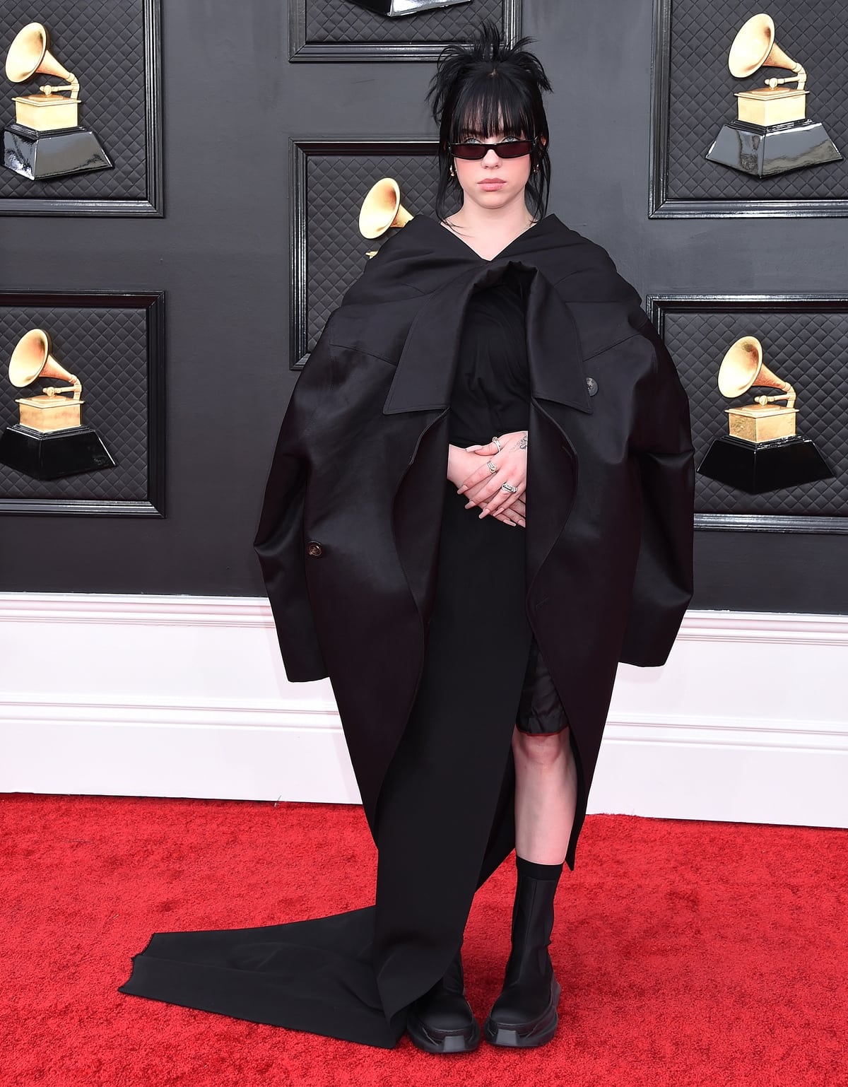 At the 64th Annual Grammy Awards at MGM Grand Garden Arena on April 3, 2022, in Las Vegas, Billie Eilish embraced Rick Owens’ signature dark minimalism in a sculptural, floor-length black ensemble layered under an oversized coat, grounding the dramatic silhouette with thick-soled black platform boots that added weight, height, and a utilitarian edge to the monochrome look