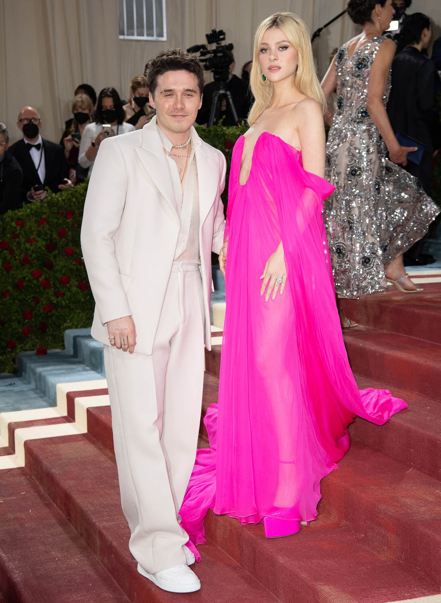 Brooklyn Beckham and Nicola Peltz turn heads at the 2022 Met Gala, with Brooklyn in a relaxed cream Valentino suit and sheer shirt, while Nicola stuns in a hot pink Valentino couture gown with sky-high platform heels and emerald drop earrings