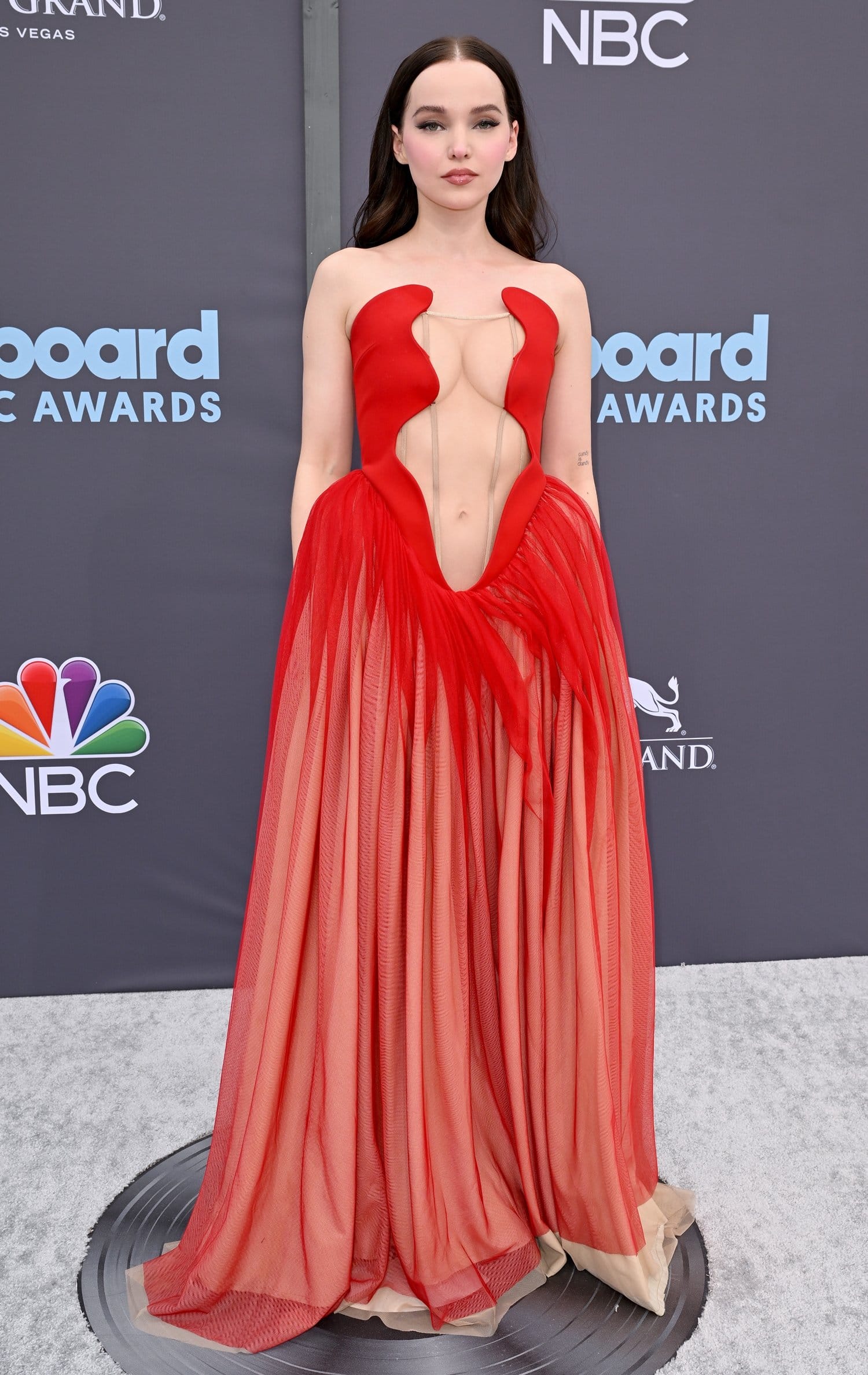 Dove Cameron commands attention in a daring red Ashlyn gown featuring sculptural cutouts and a tulle-and-mesh skirt, paired with black Giuseppe Zanotti platform heels at the 2022 Billboard Music Awards held at the MGM Grand Garden Arena on May 15, 2022, in Las Vegas