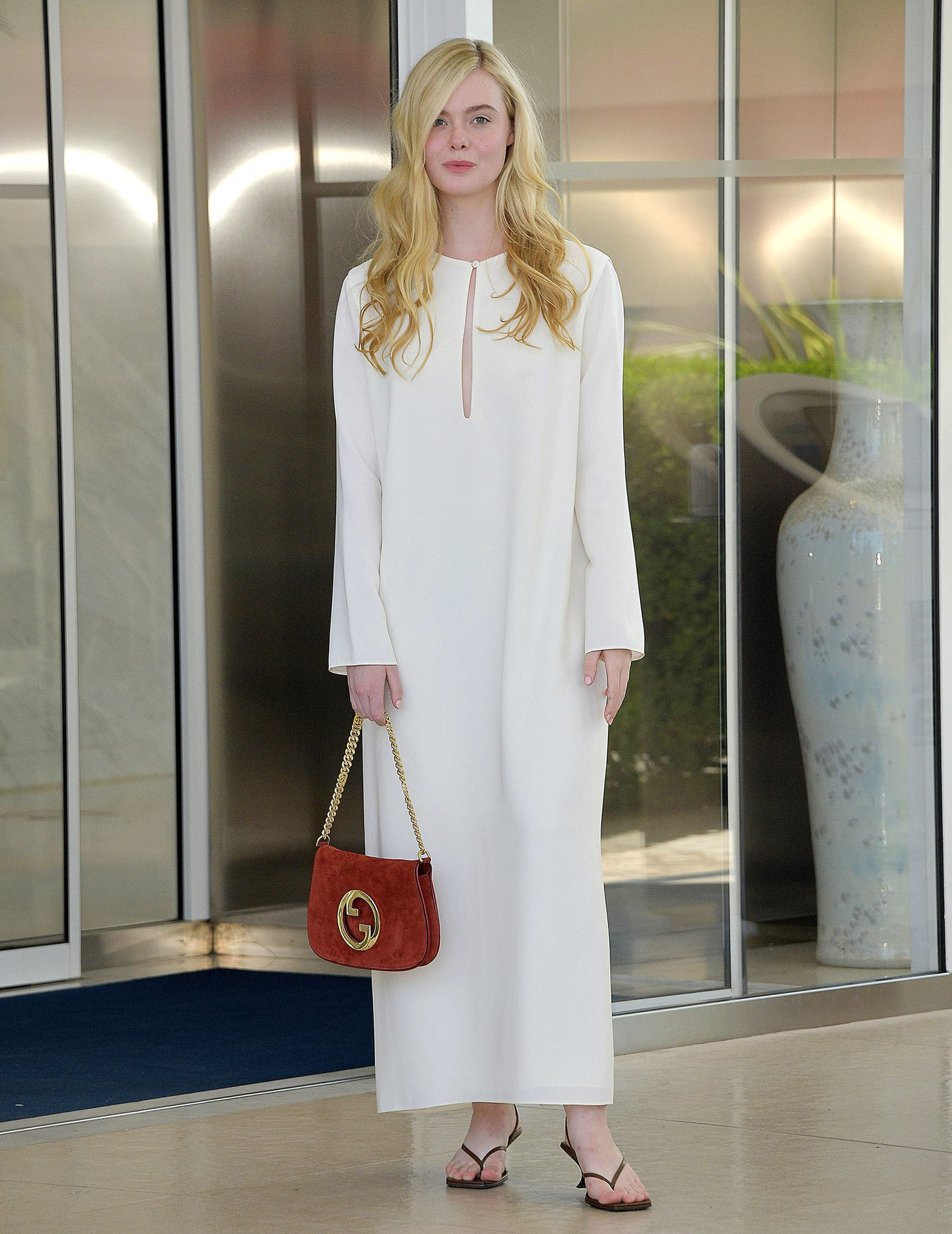 Elle Fanning steps out of the Hôtel Martinez on May 19, 2022, wearing The Row’s white “Ellita” column dress, styled with minimalist thong sandals and a red Gucci bag for an effortlessly polished Cannes off-duty look