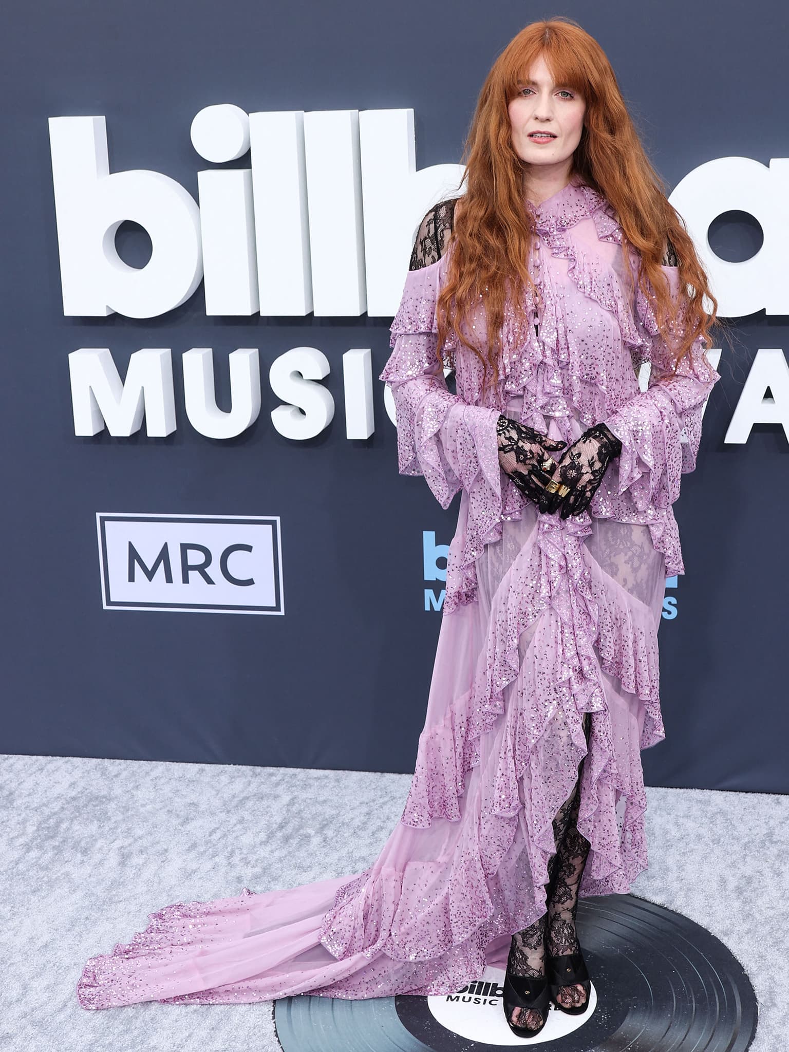 The flowing Gucci design reveals sheer lace panels beneath the ruffled lavender fabric, creating a theatrical silhouette that balances Florence Welch’s bohemian styling with sharp black accessories and platform mules