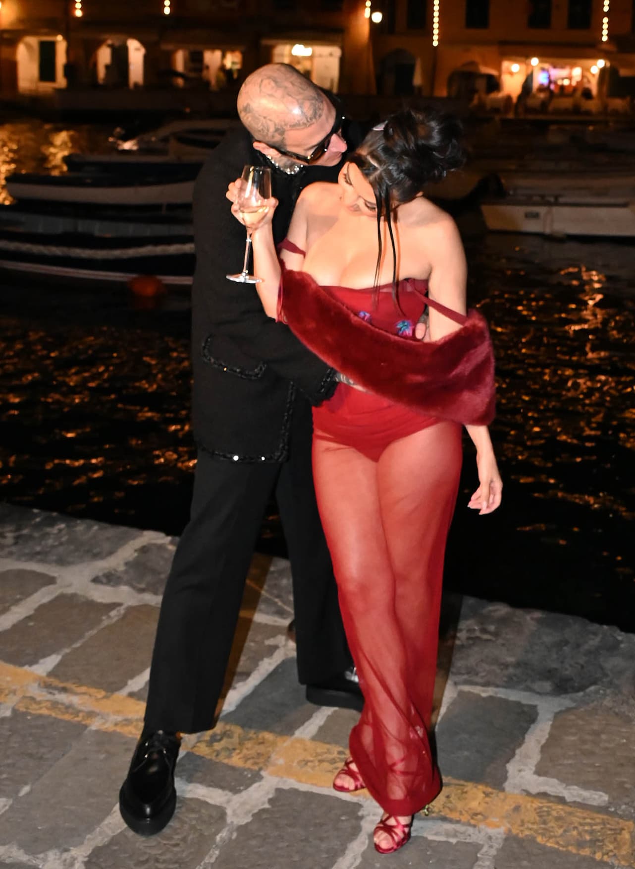 Kourtney Kardashian turns heads in a sheer red Dolce Gabbana Spring 1998 dress paired with jeweled satin rose-heel sandals as Travis Barker embraces her during their pre-wedding dinner at Ristorante Puny in Portofino on May 20, 2022