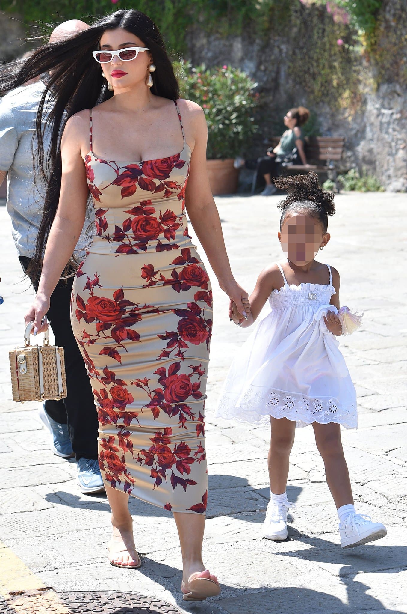 Kylie Jenner strolls through Portofino in a figure-hugging ’90s Dolce Gabbana rose-print dress styled with white cat-eye sunglasses and transparent Gianvito Rossi “Metropolis” Plexi-strap sandals while holding hands with daughter Stormi