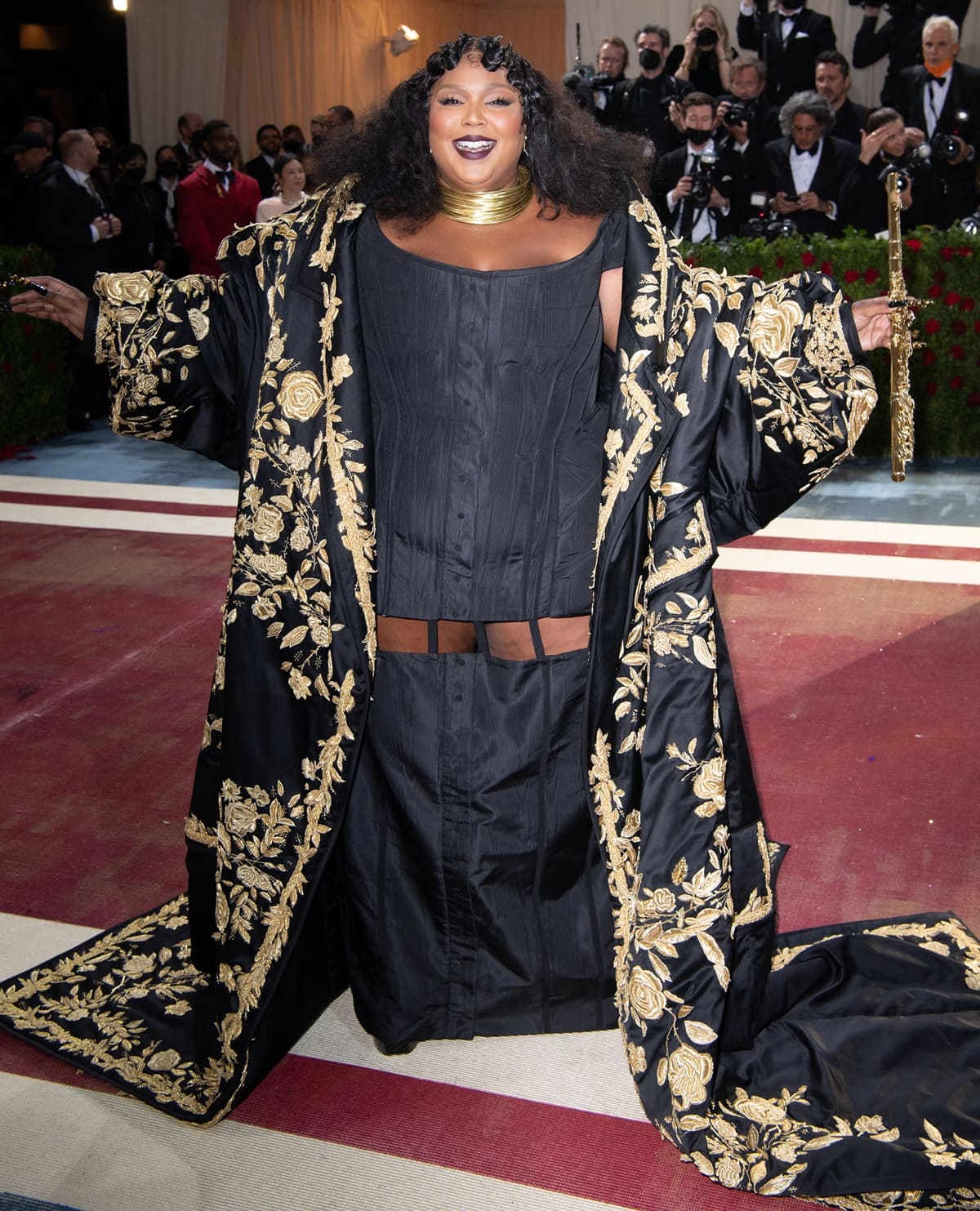 Lizzo wearing a custom Thom Browne black corseted dress beneath a hand-embroidered black-and-gold floral coat, accessorized with a stacked Lorraine West gold choker and an 18K gold John Lunn flute