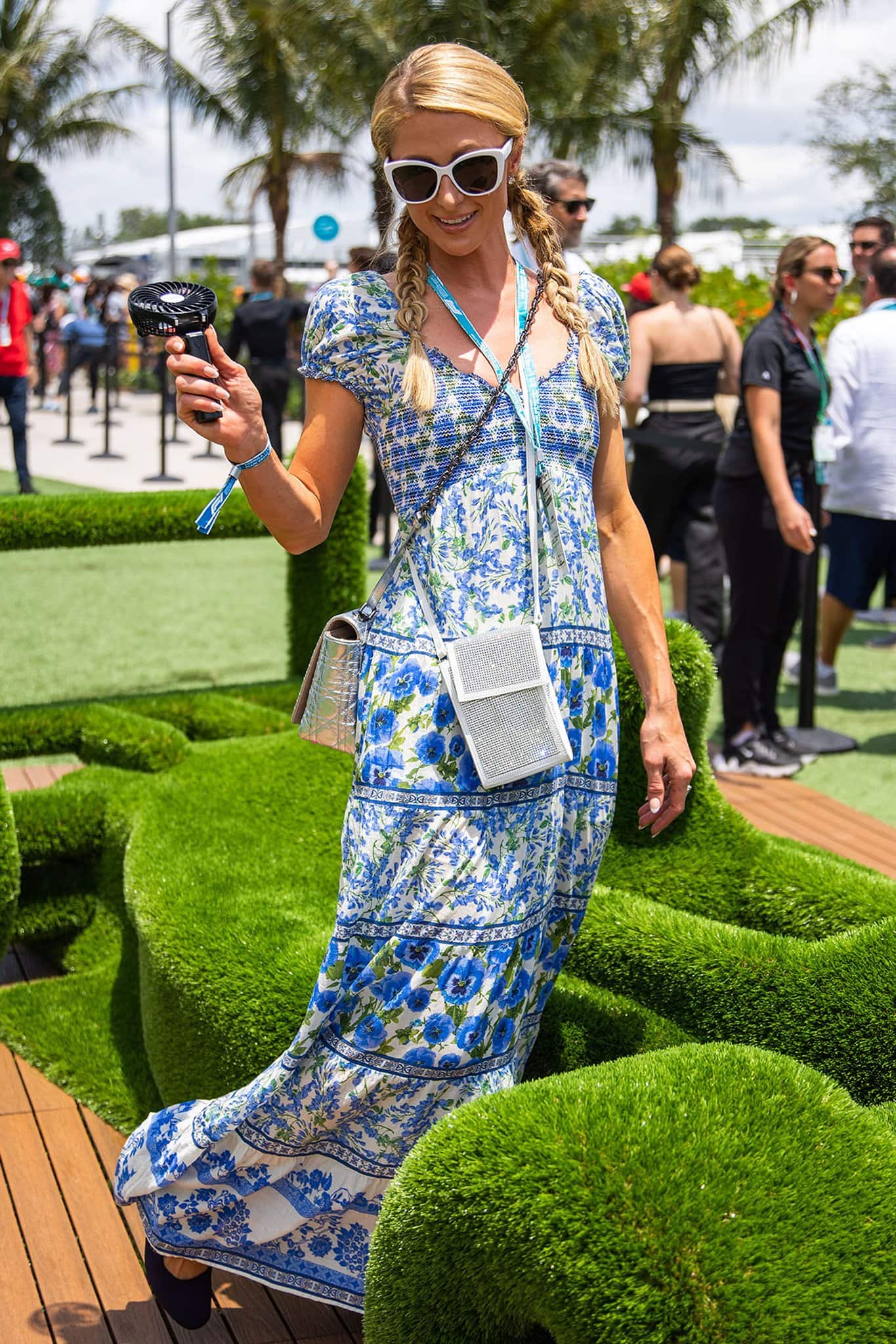 Paris Hilton embraces effortless Grand Prix glamour in Alice + Olivia’s floral-print “Rosalyn” maxi dress, pairing the smocked blue-and-white gown with oversized white sunglasses, braided pigtails, a metallic Chanel crossbody bag, and navy pointed-toe flats for a polished yet playful trackside look at the Miami International Autodrome