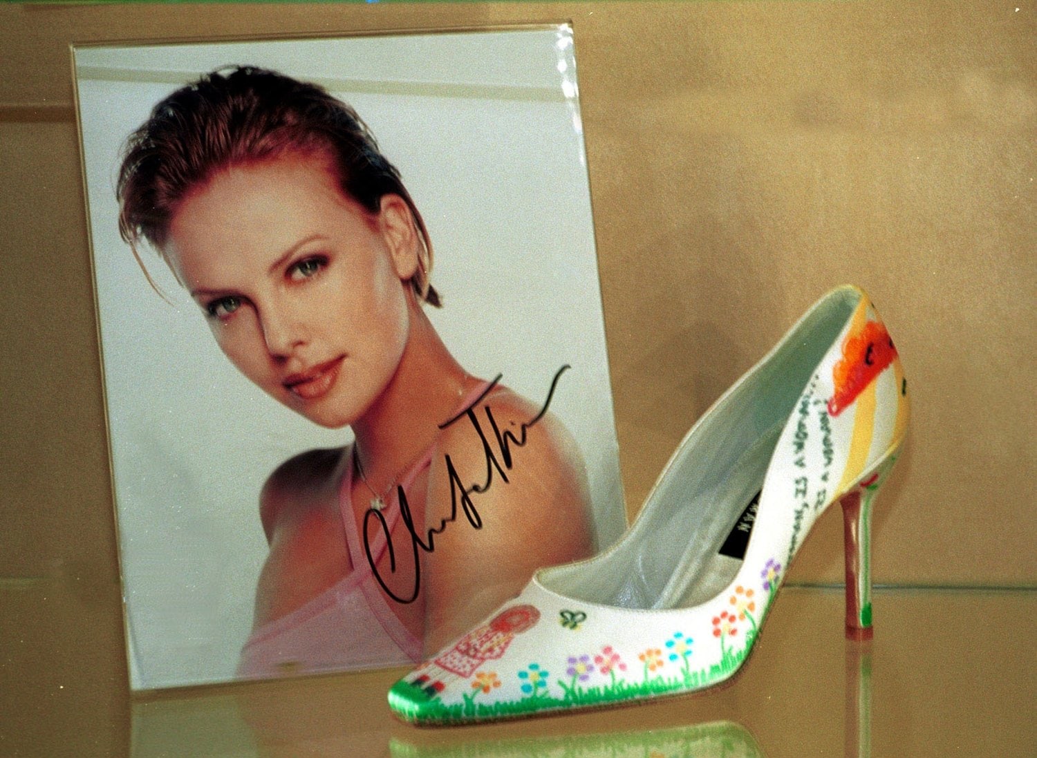 Charlize Theron’s hand-painted Stuart Weitzman pump, customized for charity and displayed alongside her signed portrait — a fusion of artistry, philanthropy, and timeless shoe design