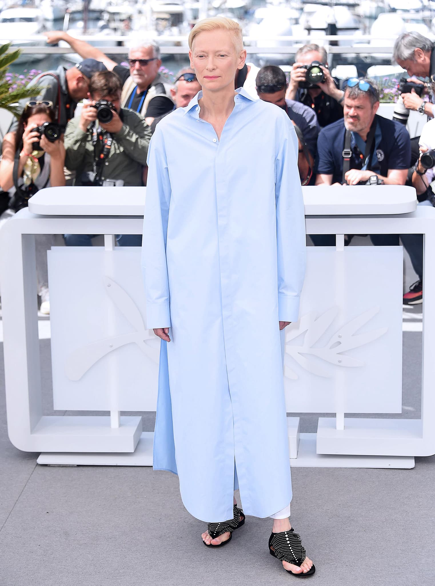 Swinton shifts from red-carpet glamour to relaxed proportions in an oversized pale-blue Alaïa shirt dress, white trousers, and heavily studded flat sandals